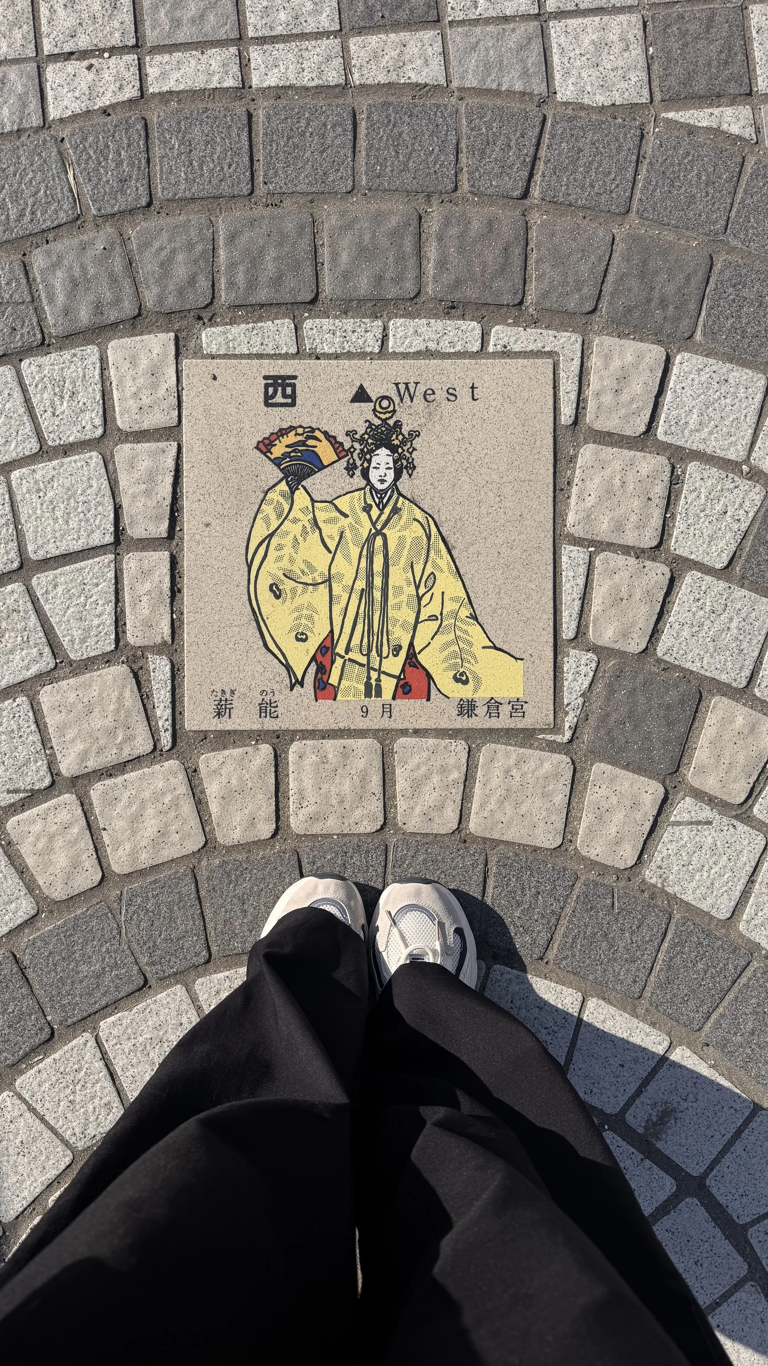 Pessoa de tênis preto e branco olhando para uma placa de rua de mosaico que mostra uma figura histórica japonesa vestida de amarelo, segurando um leque, com texto em inglês 'West' e caracteres chineses.