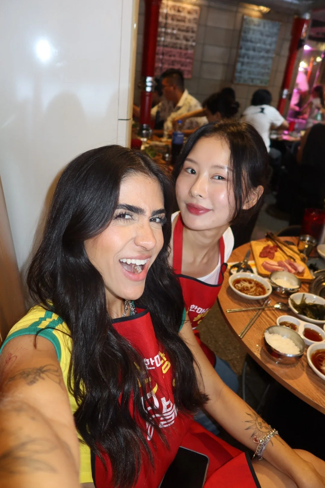 Duas mulheres sorrindo para a câmera em um restaurante com comida coreana na mesa.