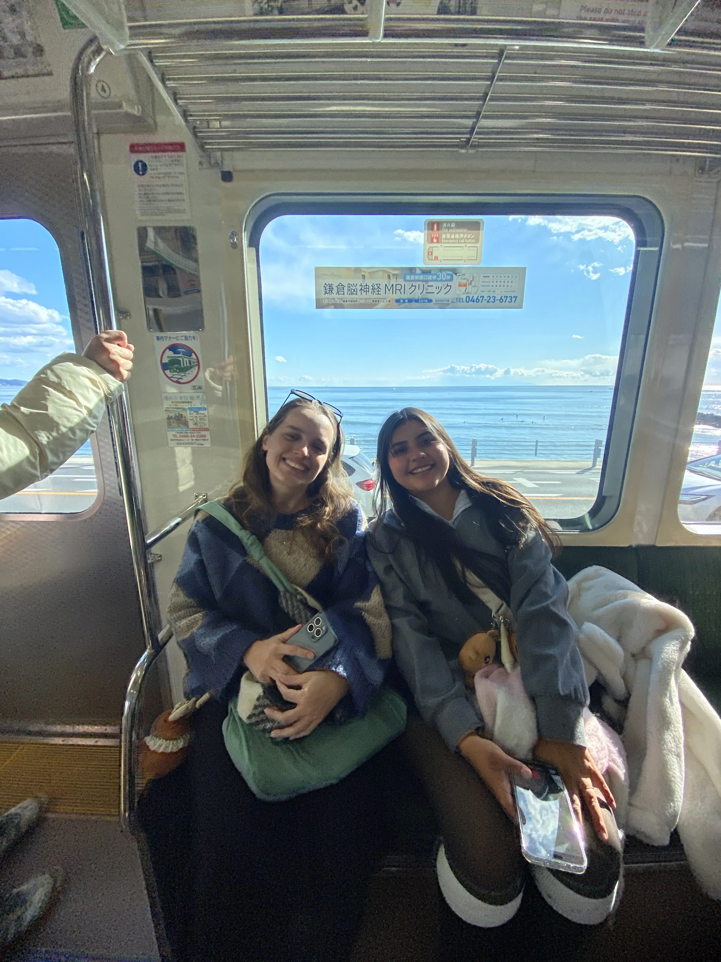Duas jovens mulheres sentadas em um transporte, sorrindo para a câmera, com uma vista do oceano ao fundo através de uma janela grande, ao lado de uma mochila e um cachorro de pelúcia.