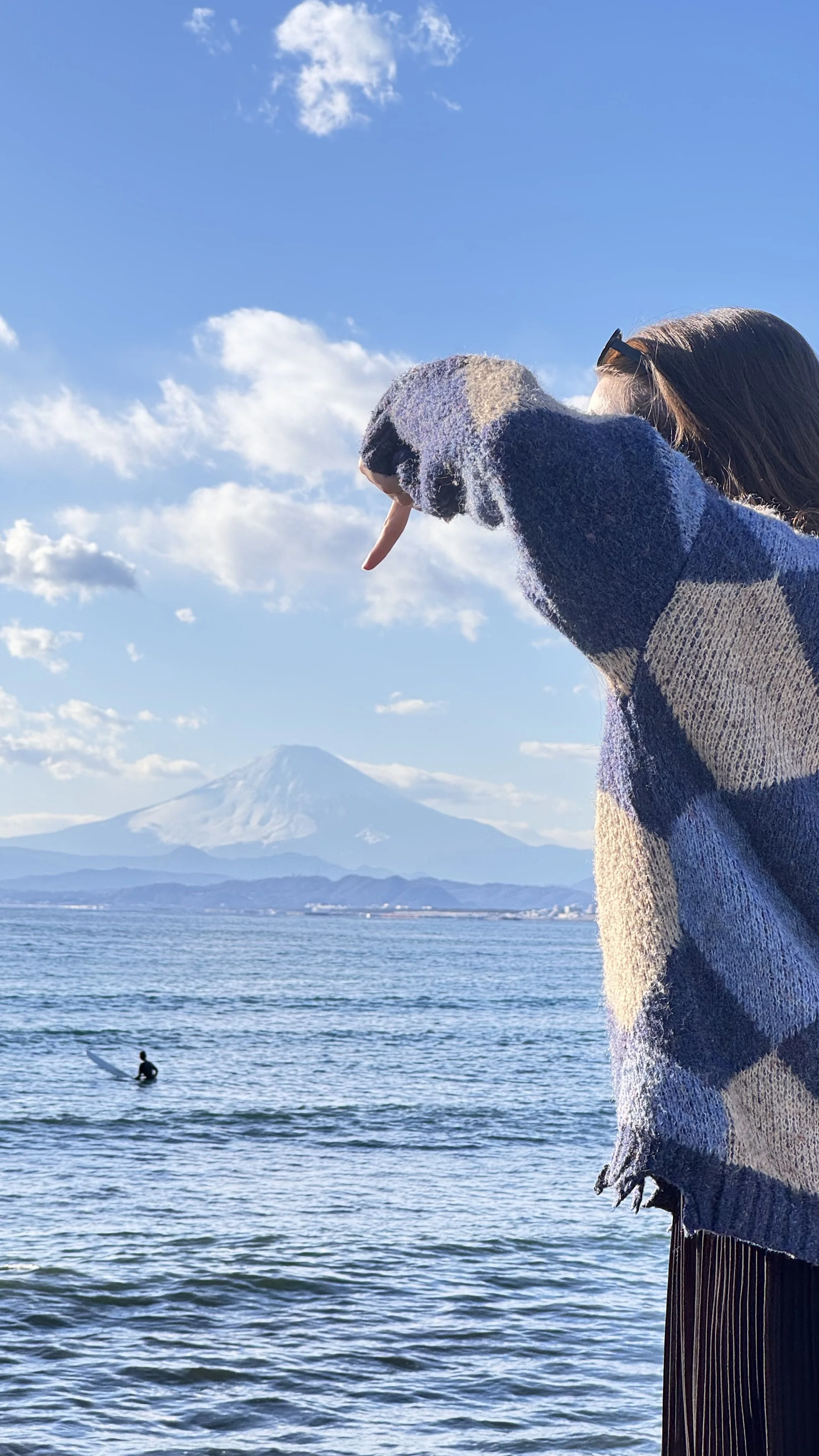 Pessoa vestida com suéter listrado apontando para o Monte Fuji ao longe enquanto está na costa de um lago com um surfista ao fundo.
