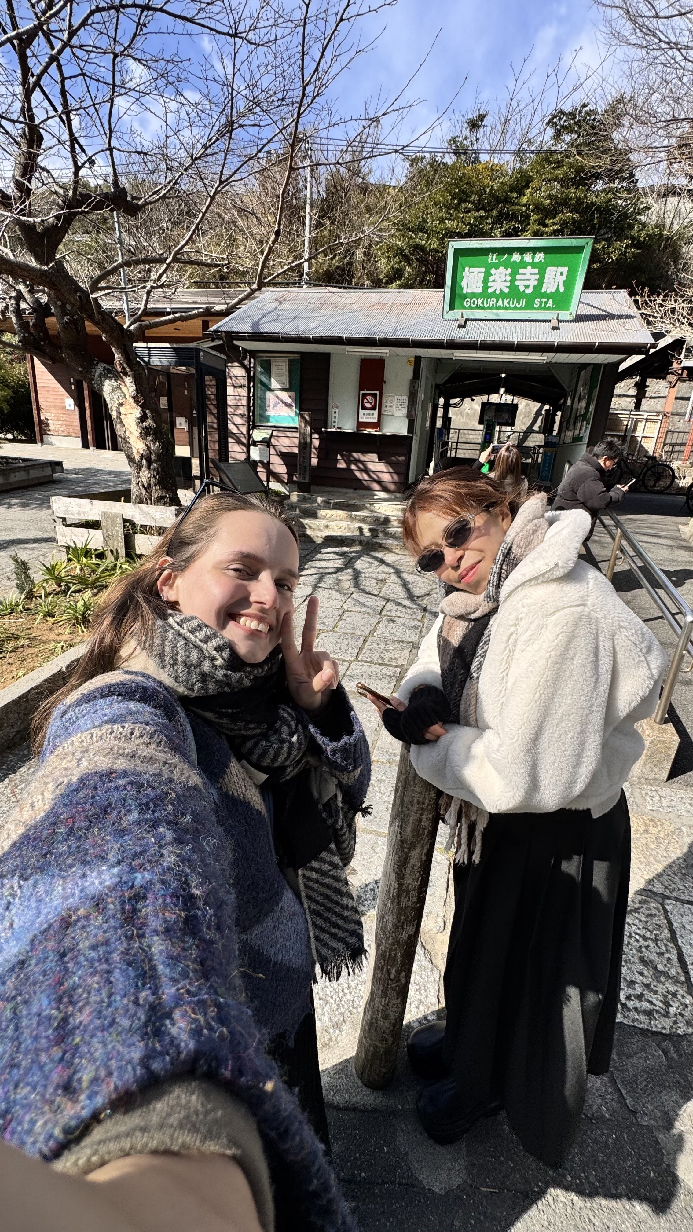 Duas mulheres sorrindo na frente da estação de trem Gokurakuji, em um dia ensolarado, com árvores sem folhas ao fundo.