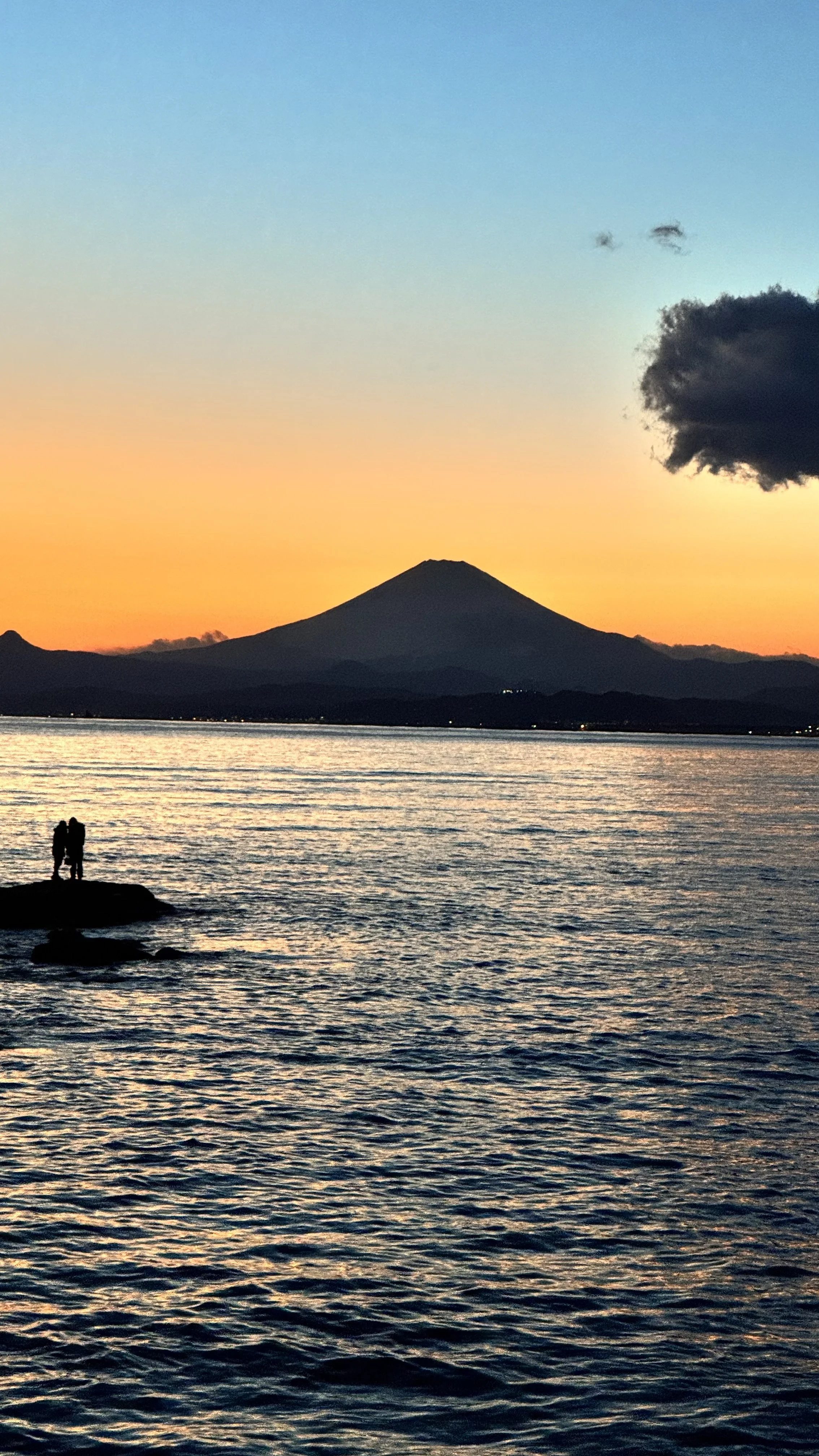 Pôr do sol com vista de um lago ou mar, uma silhueta de duas pessoas em uma pedra na água, ao fundo um vulcão.