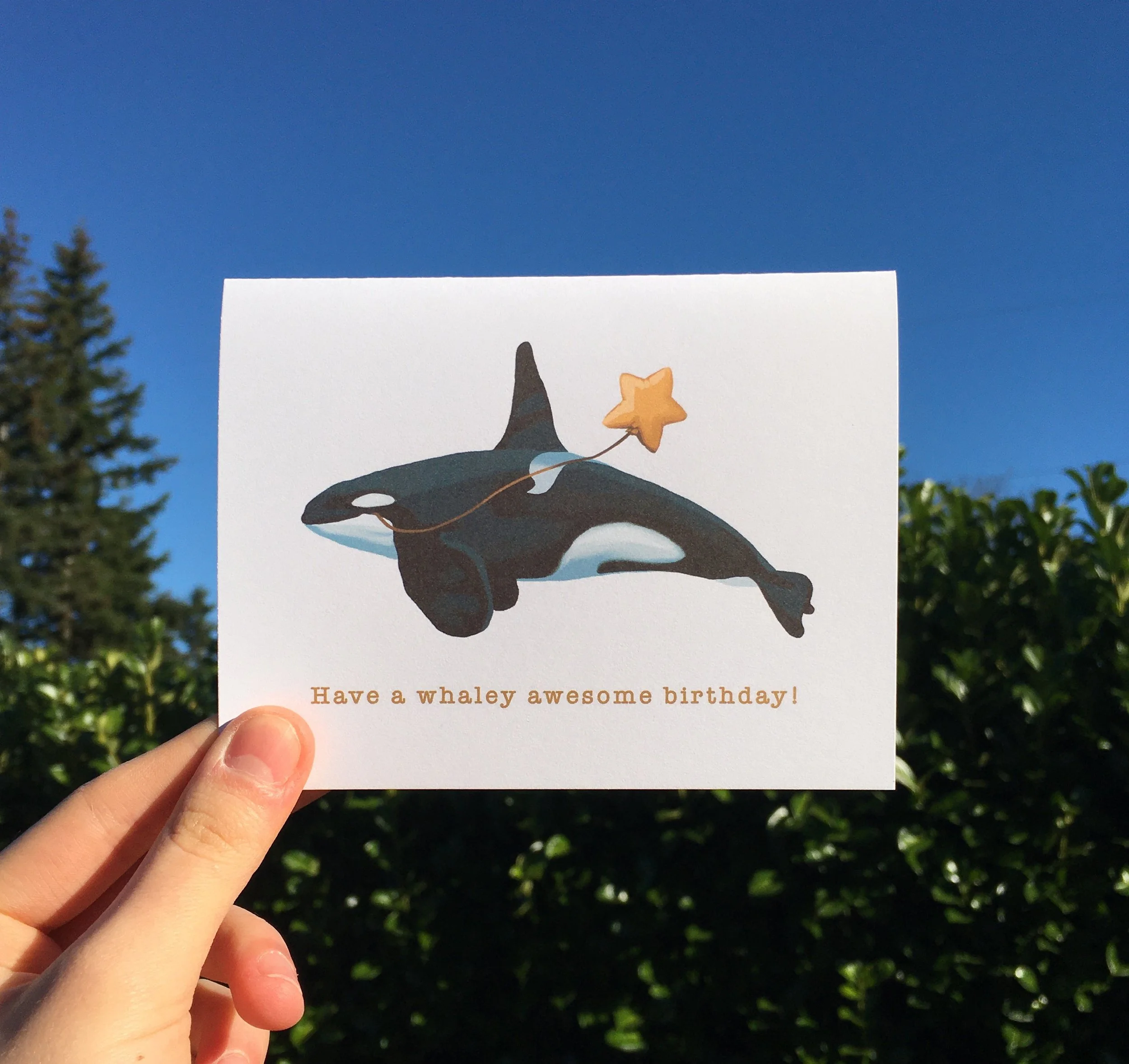 A birthday card featuring an orca whale holding a star-shaped balloon with the message "Have a whaley awesome birthday!" against a backdrop of clear blue sky and green trees.