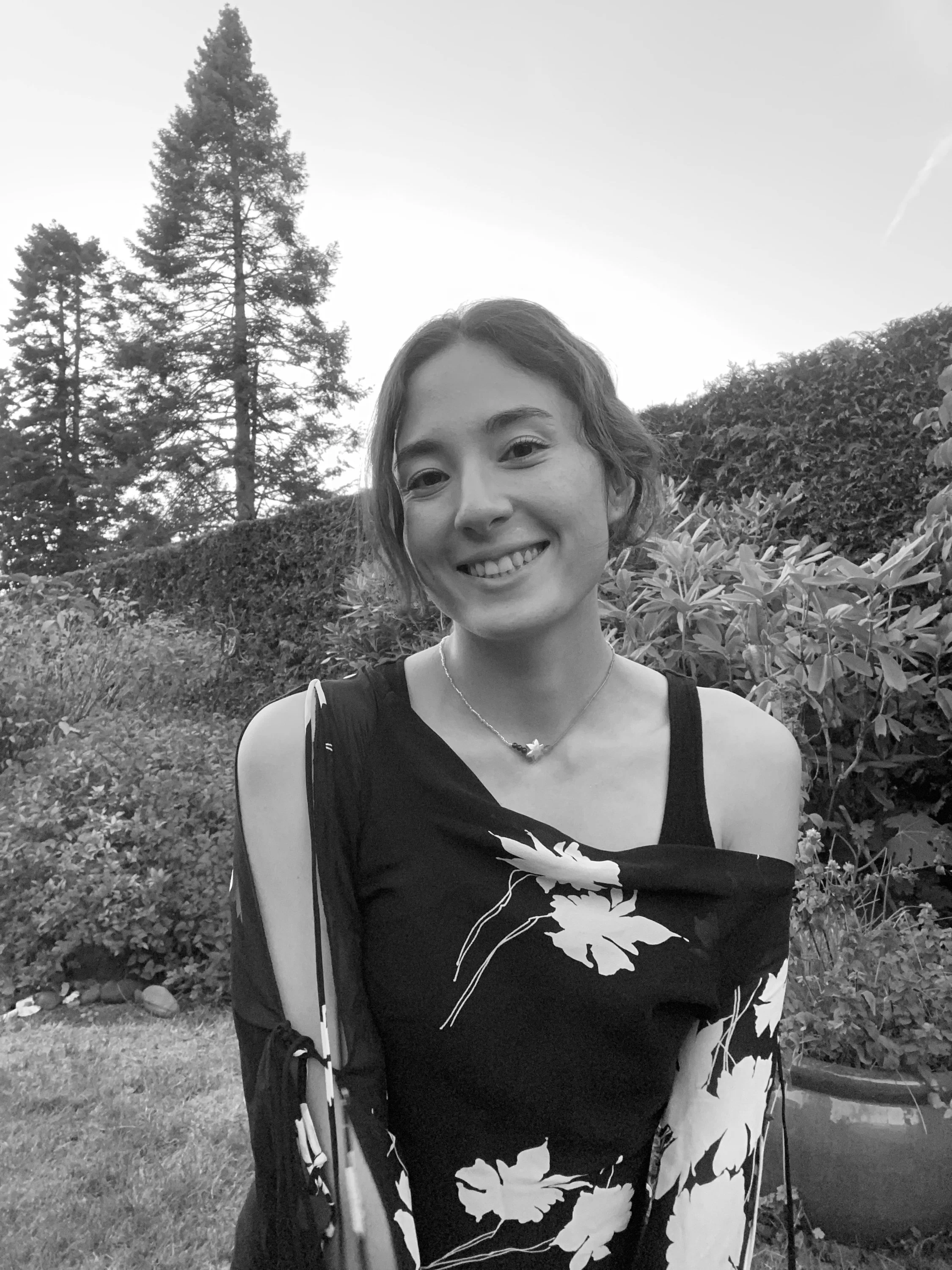 A young woman with short, wavy hair smiling outdoors, wearing a floral dress and a necklace with a star pendant, standing in a garden with trees and bushes in the background.