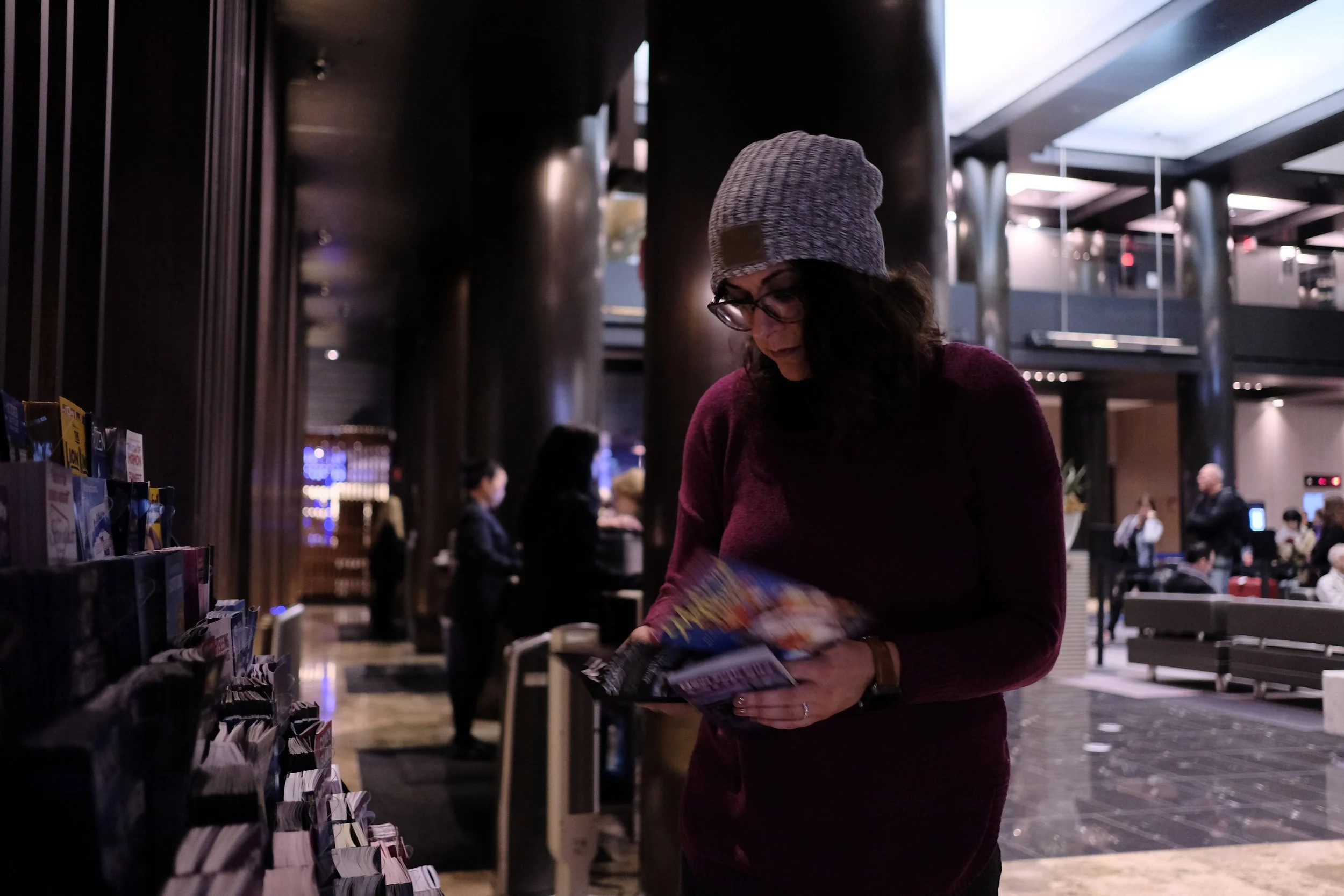 A woman with glasses and a gray beanie browsing magazines in a lobby or reception area with modern decor and other people in the background.