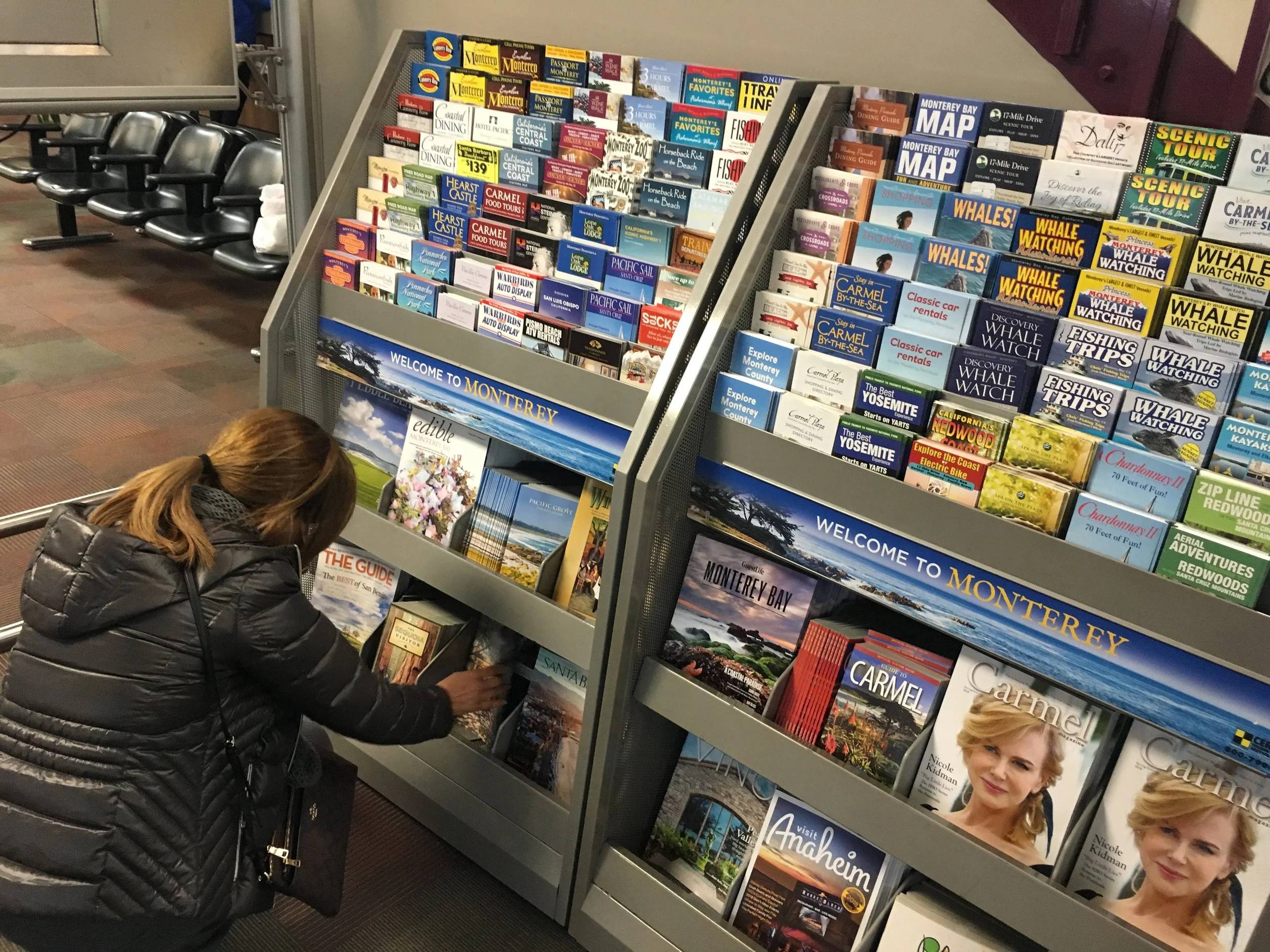 A woman with brown hair, wearing a black puffer jacket, examining travel brochures at a rack in an airport or visitor center.