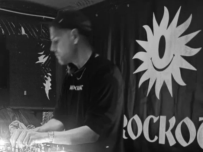 A DJ wearing a black shirt, standing behind DJ equipment, in a club with a black curtain backdrop featuring a smiling sun logo and the word "ROCKO".