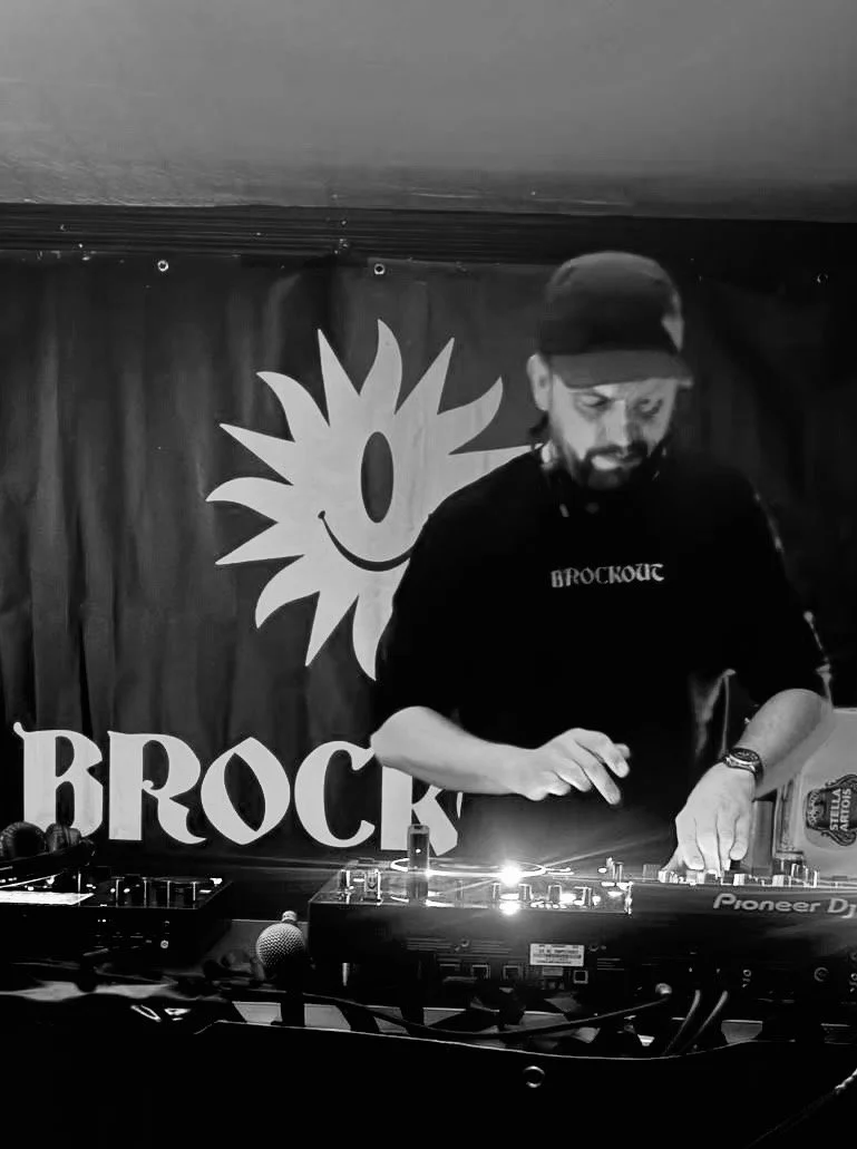A man DJing at a booth with a smiling sun logo behind him and the word 'BROCK' visible.