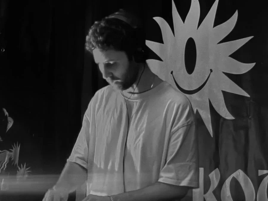 A DJ with curly hair wearing a white T-shirt playing music with headphones on, standing in front of a backdrop with a sun-like smiling face with rays and the word "Koo" partially visible.