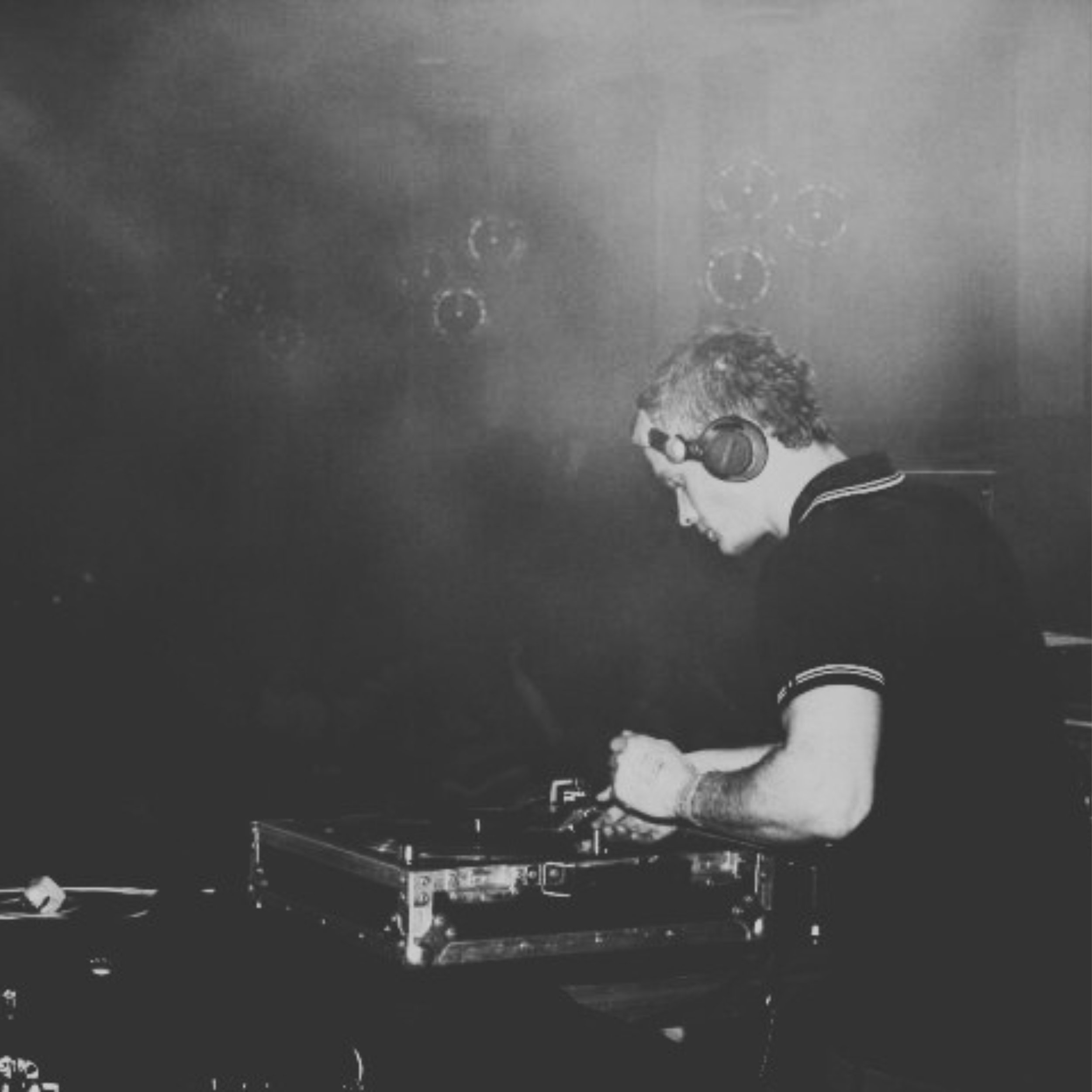 A young male DJ with curly hair wearing headphones, working with DJ equipment in a dark setting.
