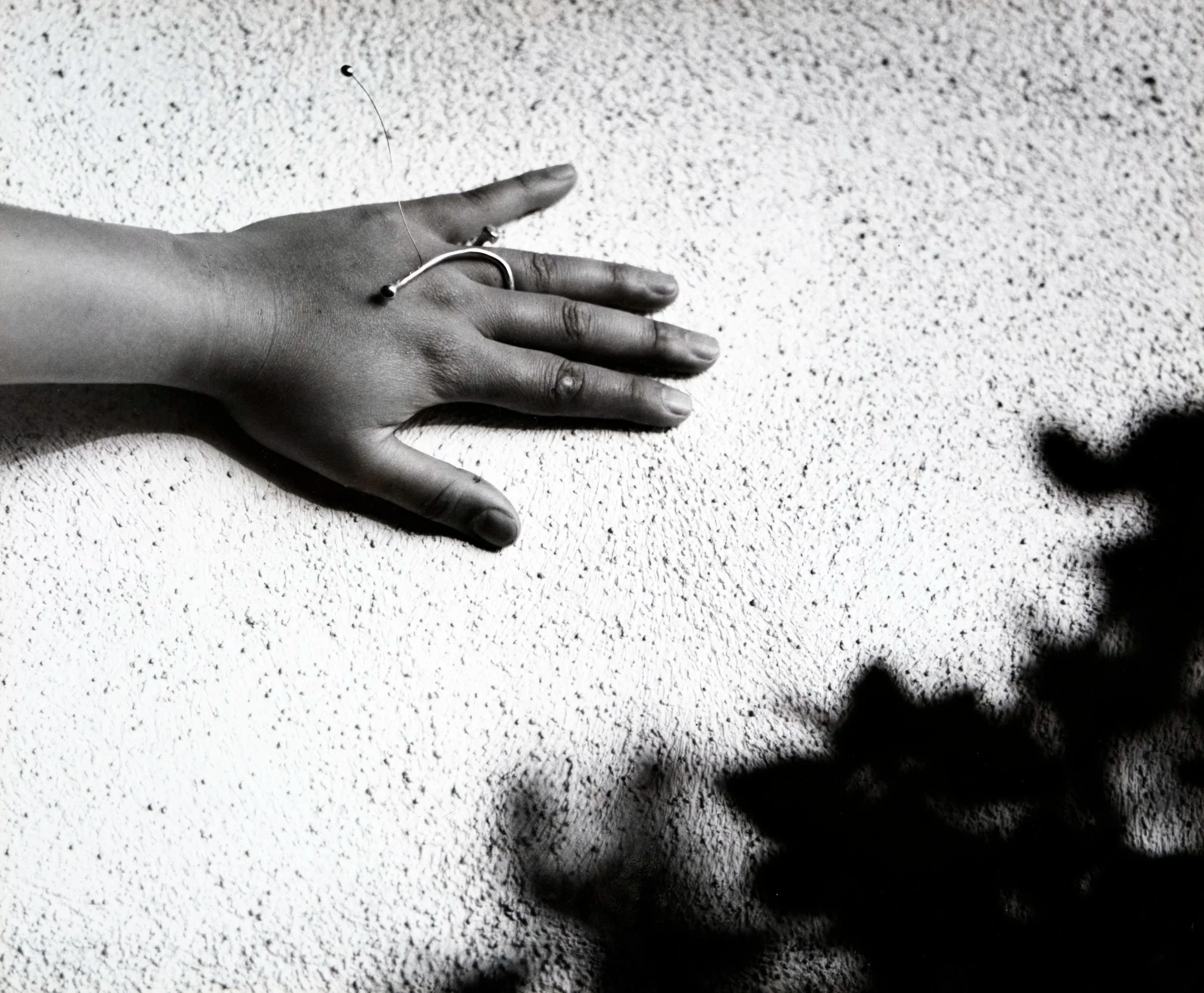 A hand wearing a ring is placed on a textured white surface, with a black string or thread and a small bead near the fingers.