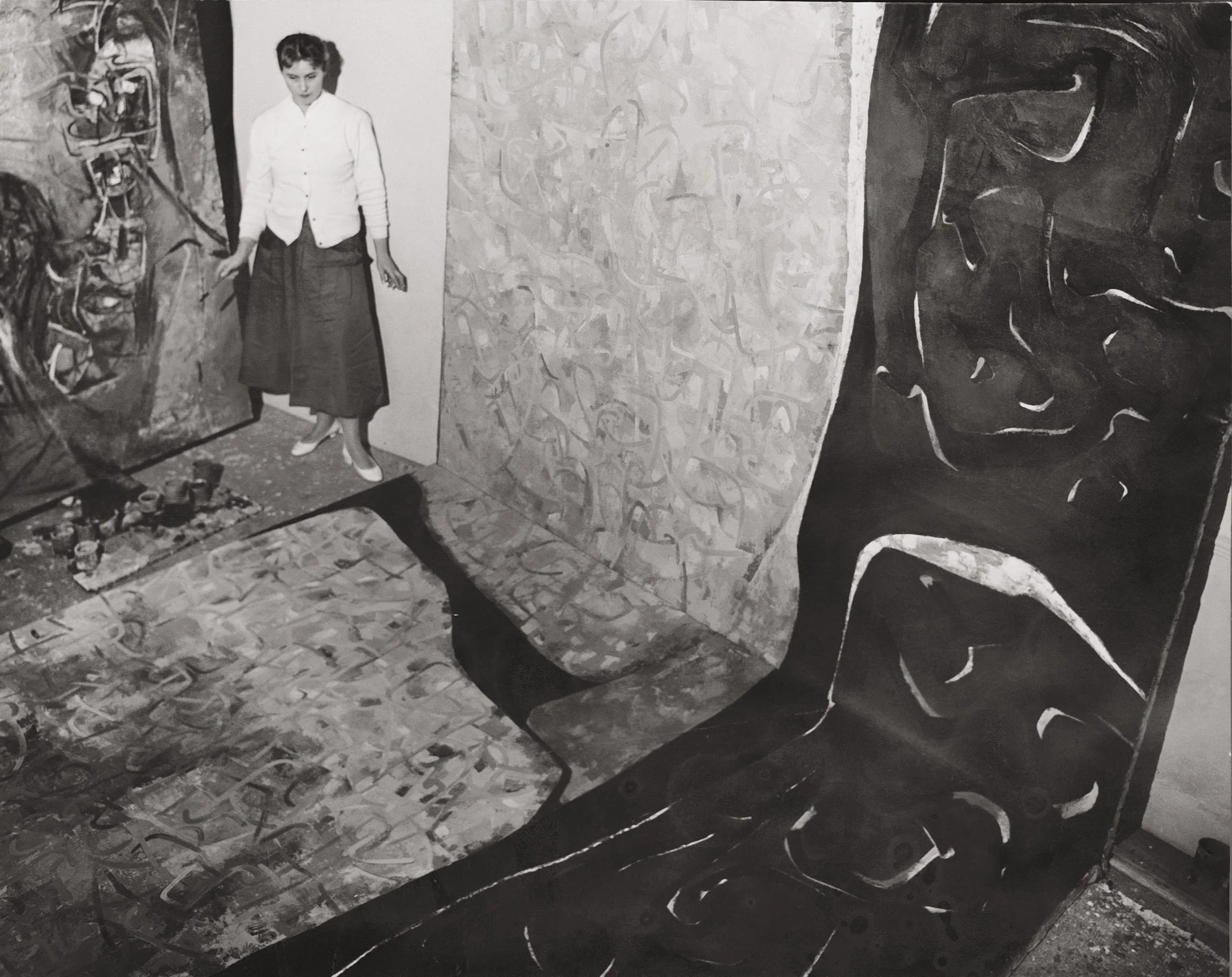 Black and white photograph of an art gallery with abstract paintings on the walls. A woman is standing and looking at the artworks, dressed in a long skirt and a light-colored sweater. The floor and walls are decorated with various patterns and textures.