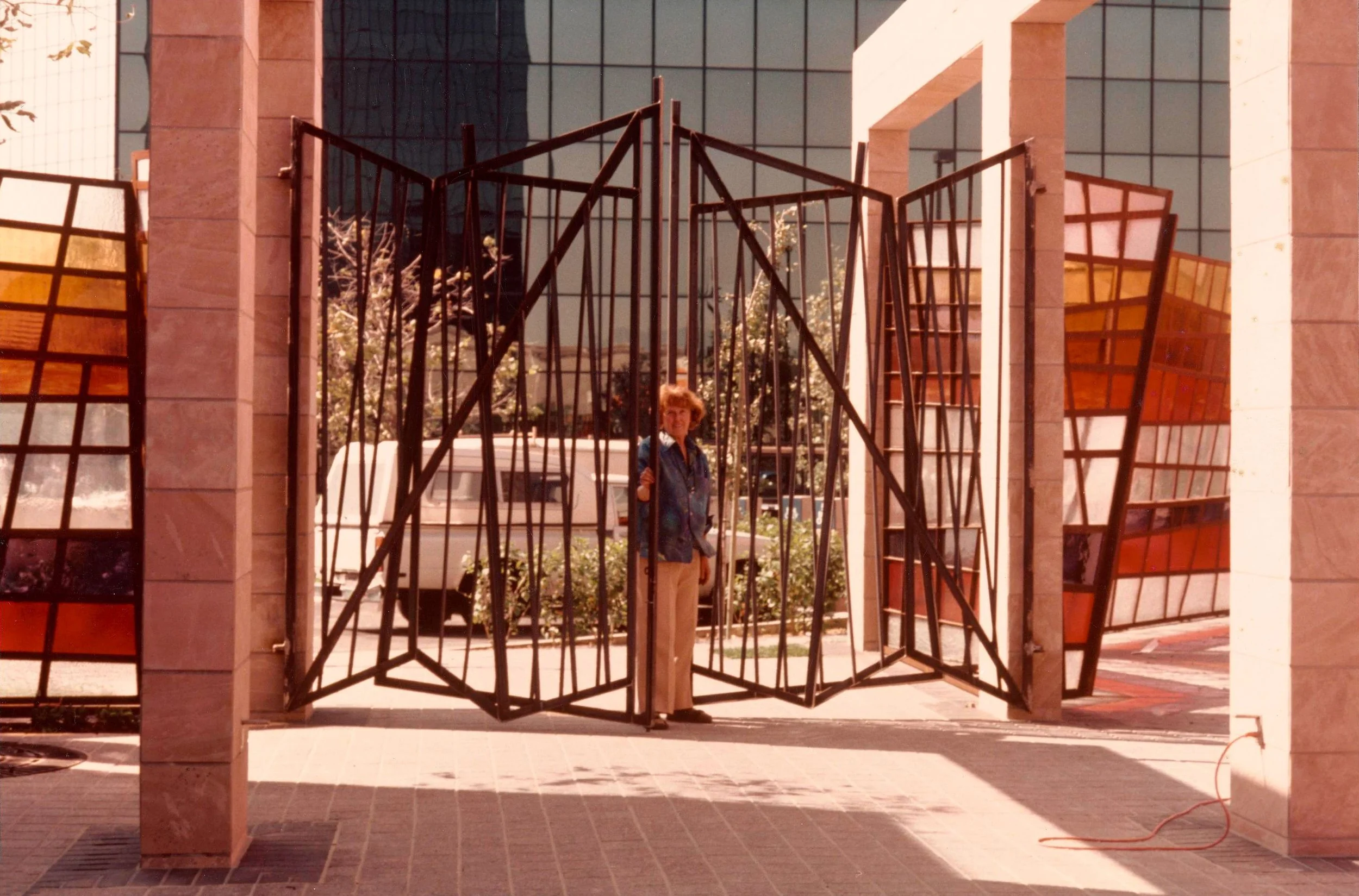 A color photograph of Claire Falkenstein standing in front of a decorative metal gate outside a building, with colored glass windows and brick pillars, in an urban setting.