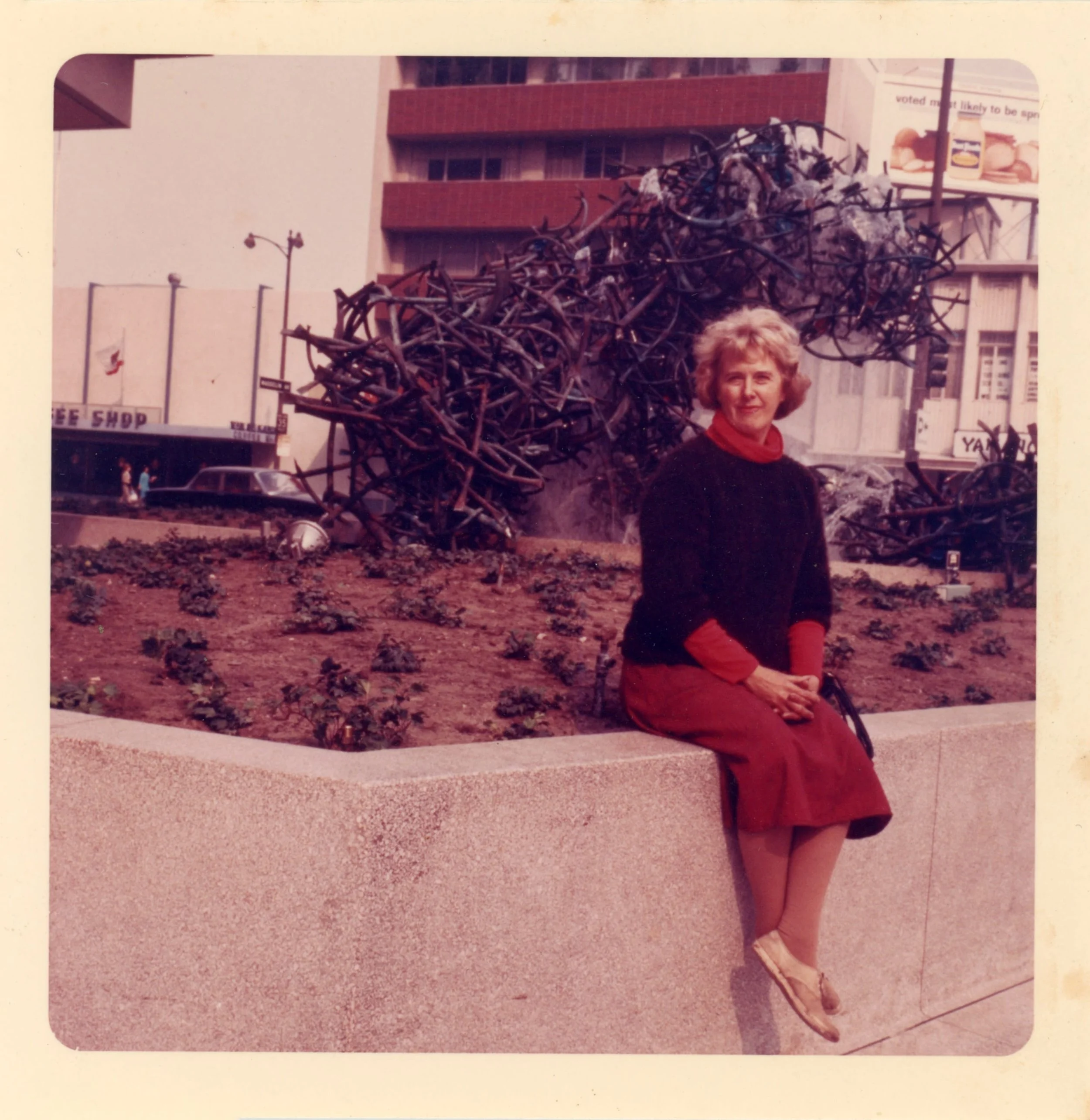 Color photograph of Claire Falkenstein seated on a concrete ledge with one arm resting on her lap, smiling at the camera. Behind her, a large fountain of metal tubing sends water cascading downward.