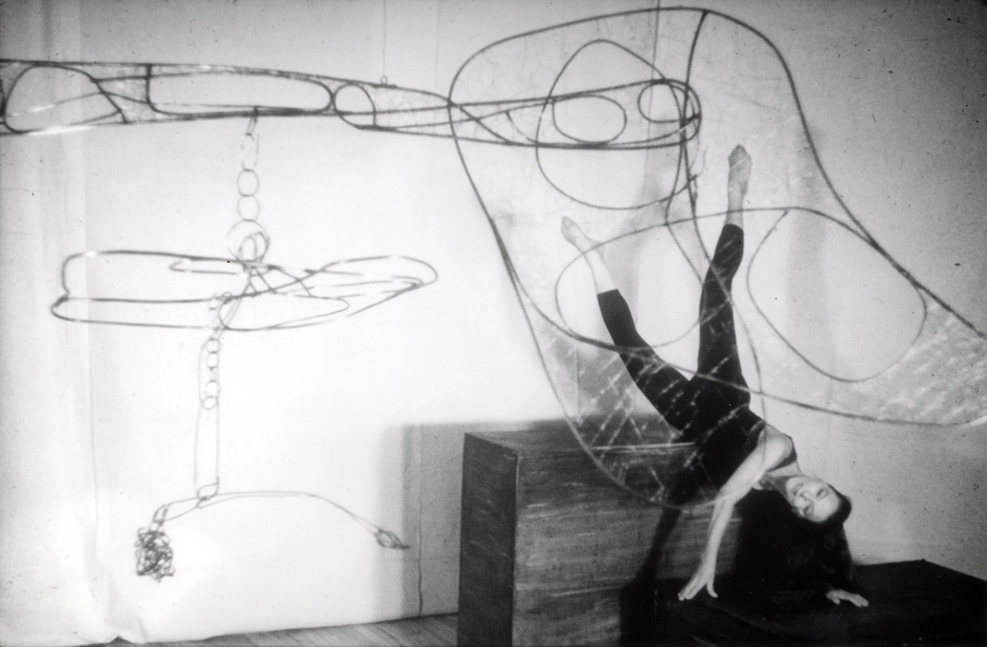 Black-and-white photograph of a suspended mobile sculpture made of metal tubing, wire, and chains. On the right, a woman appears mid-performance beneath and surrounded by the sculpture.