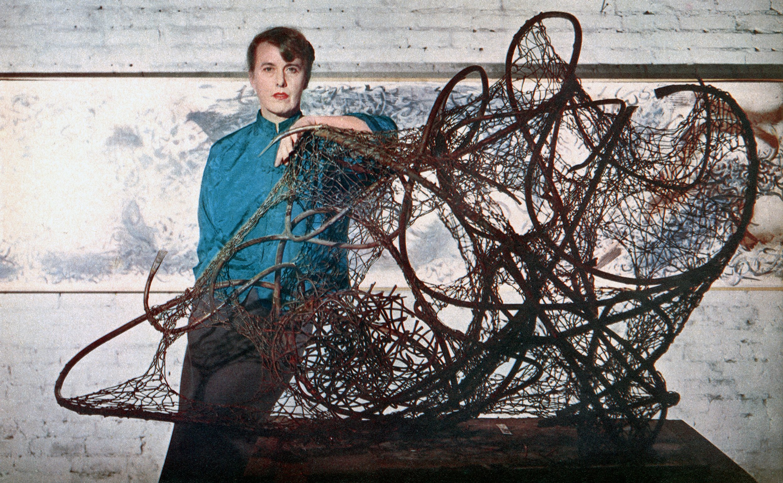 A color photograph of Claire Falkenstein standing with a large sculpture of wire and tubing in the foreground
