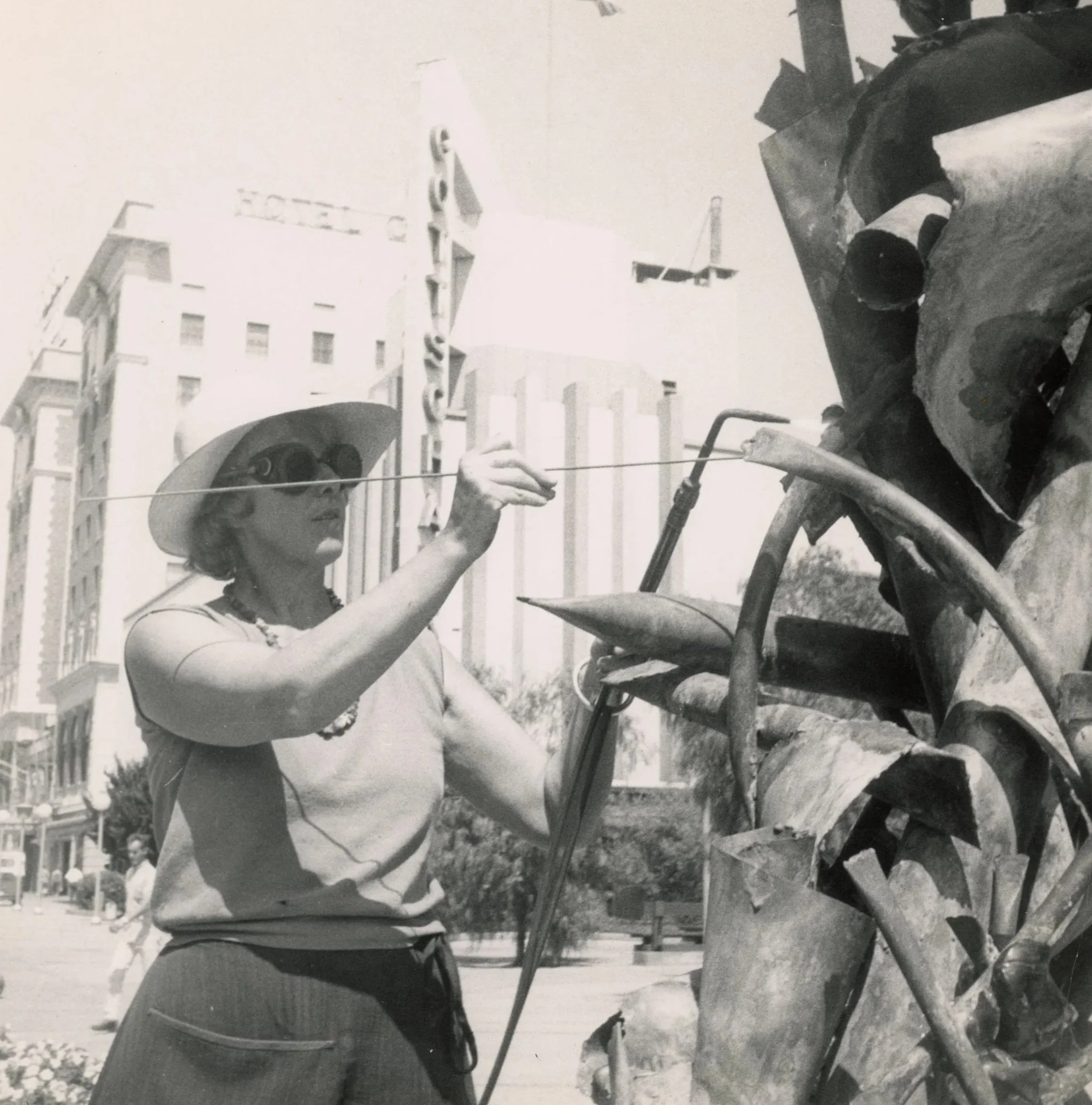 Black-and-white photograph of Claire Falkenstein wearing a hat and protective welding goggles while welding metal onto a sculpture outdoors in Fresno, California.