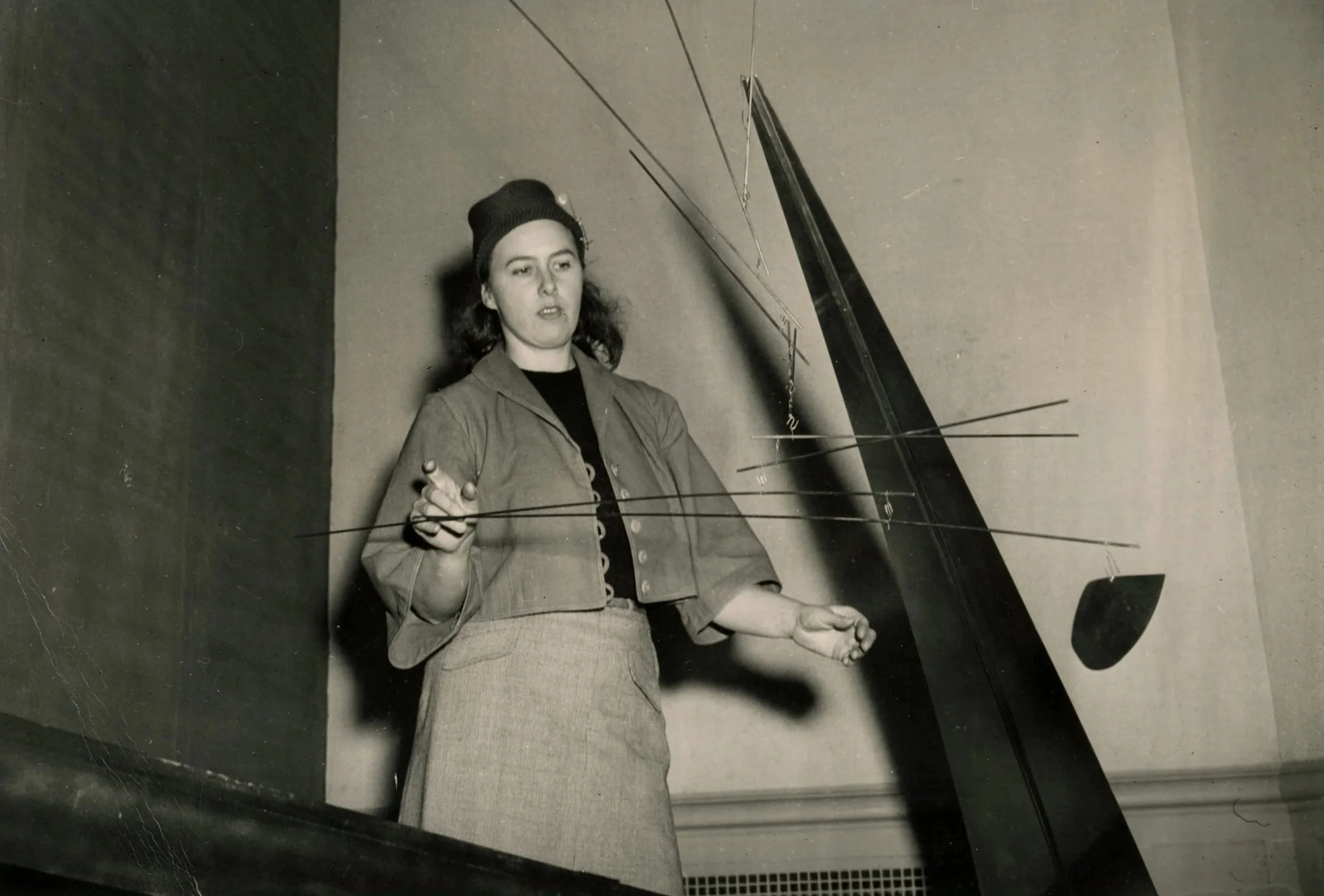 Black-and-white photograph of Claire Falkenstein standing with her mobile sculpture at the San Francisco Museum of Art, wearing a beanie, long coat, and skirt, 1948.