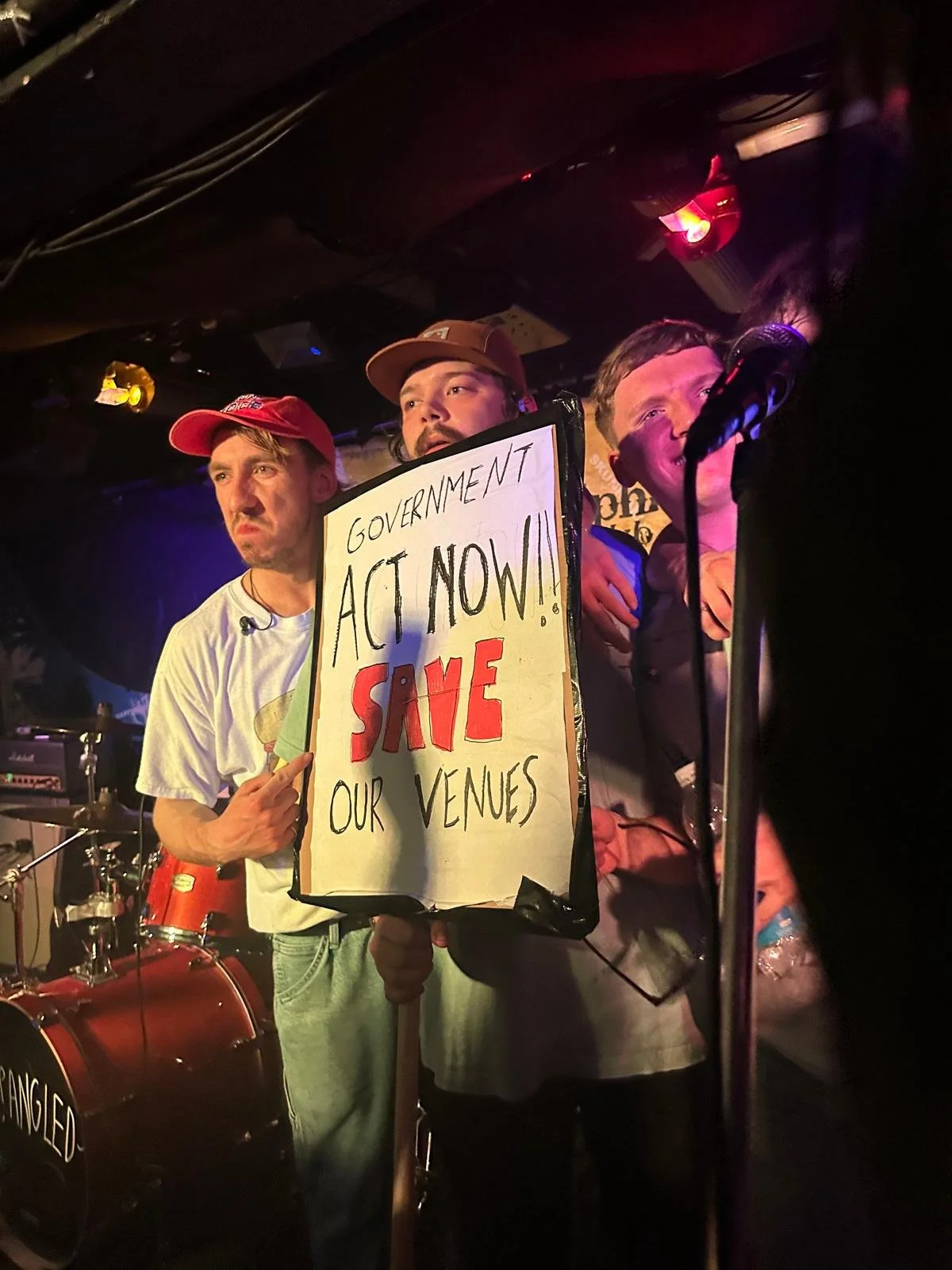 Three men in casual clothes on stage, one holding a protest sign that reads 'Government Act Now! Save Our Venues,' with a band set in the background.