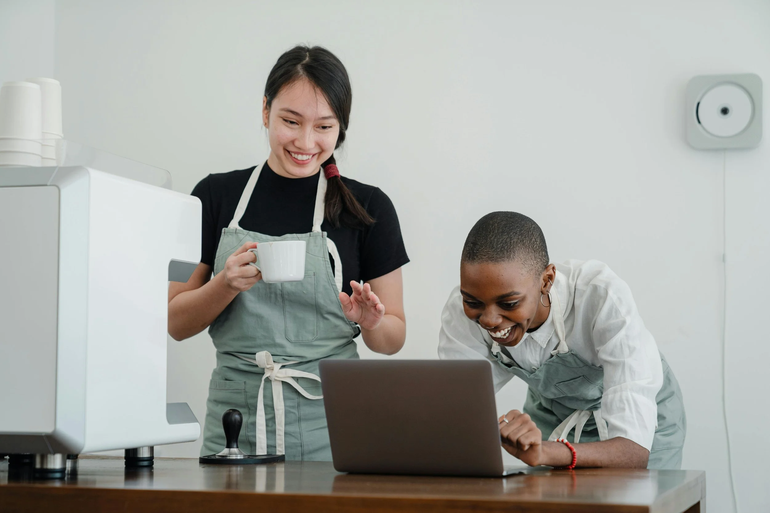 Two women in aprons, one holding a mug, looking at a laptop on a counter in a kitchen