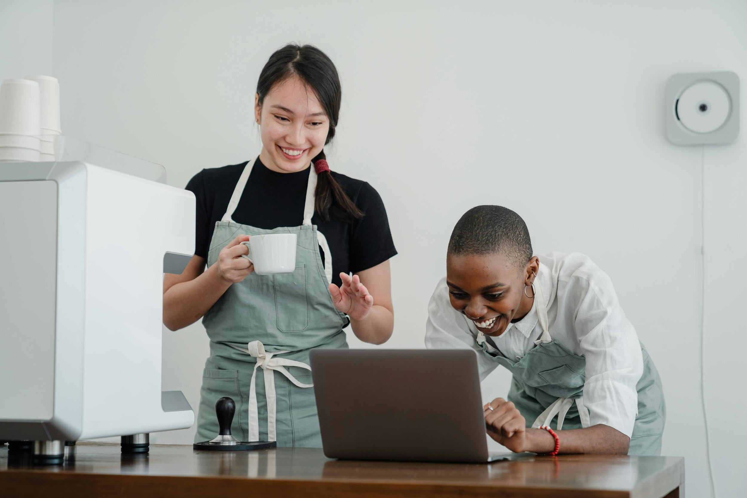 Two women, one holding a coffee mug, looking at a laptop with a smile in a kitchen