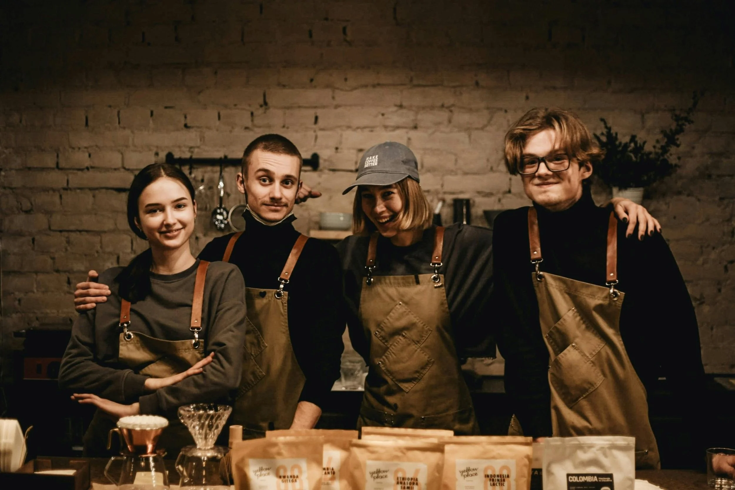 Four young adults standing together in a bakery or coffee shop, smiling and posing for the camera. They are wearing aprons, and the background shows a brick wall with shelves of kitchen items