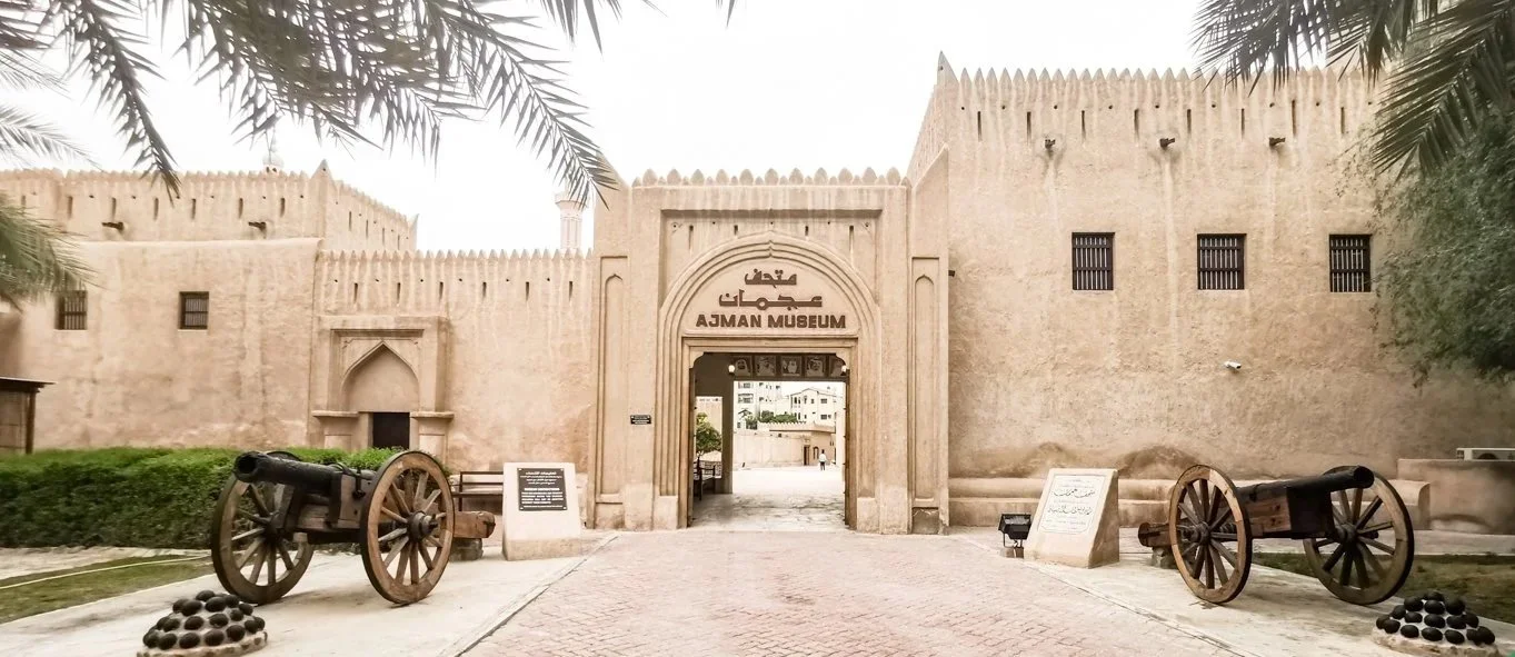 Entrance to Ajman Museum with old cannons and cannonsballs on the ground, surrounded by palm trees.