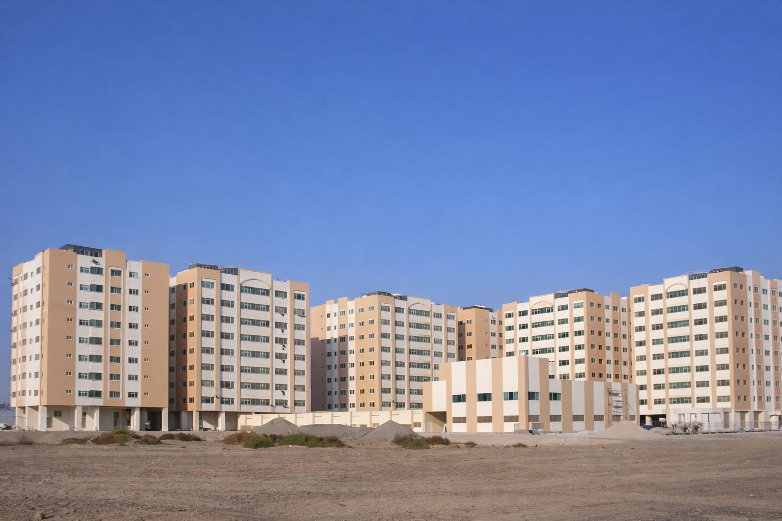 Multiple beige and white multi-story apartment buildings under a clear blue sky, with a dirt field in the foreground.