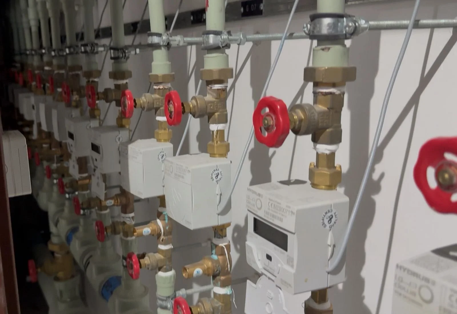 A close-up of a plumbing system with multiple pipes, valves, and meters. The valves have red handles, and the pipes are connected to white digital meters attached to the wall.