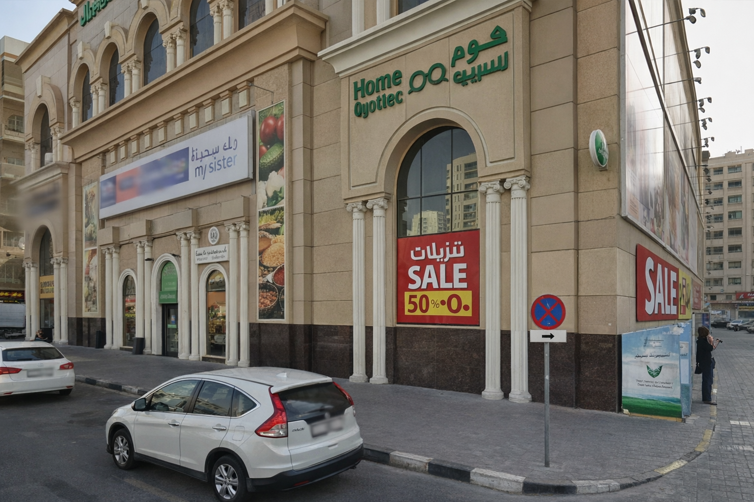 A beige commercial building with Arabic and English signage, including a large sale sign indicating 50% off. The building has Italian-style columns at the entrance, and there are a few cars parked on the street in front. A woman is standing on the sidewalk talking on her phone.