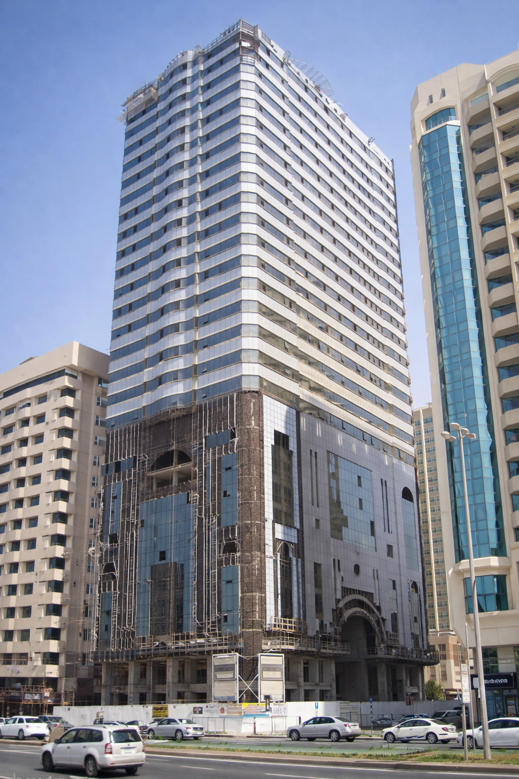 A tall modern building under construction with scaffolding at the bottom, surrounded by other high-rise buildings on a city street.