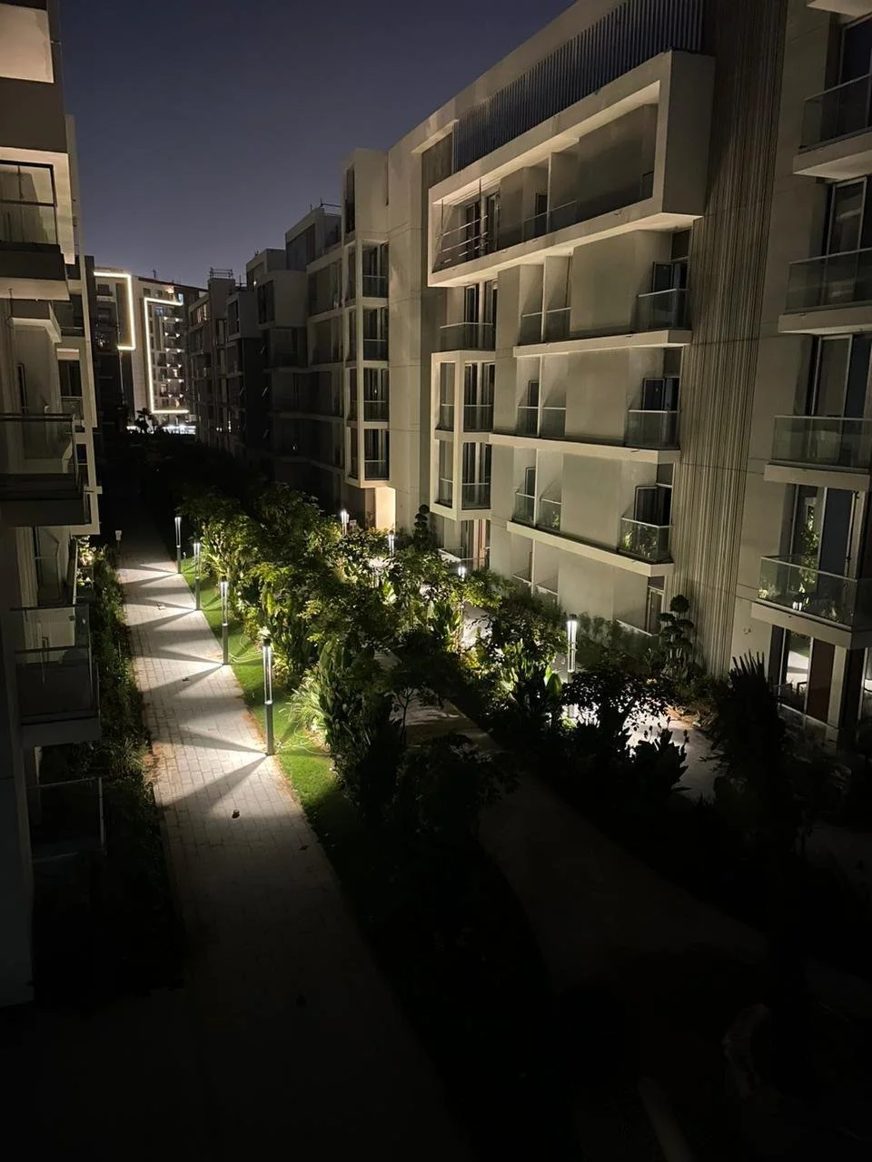 Night view of an apartment complex courtyard with illuminated walkways, lush greenery, and modern buildings with balconies.