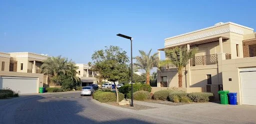 Residential street with modern beige buildings, palm trees, and parked cars under a clear blue sky.
