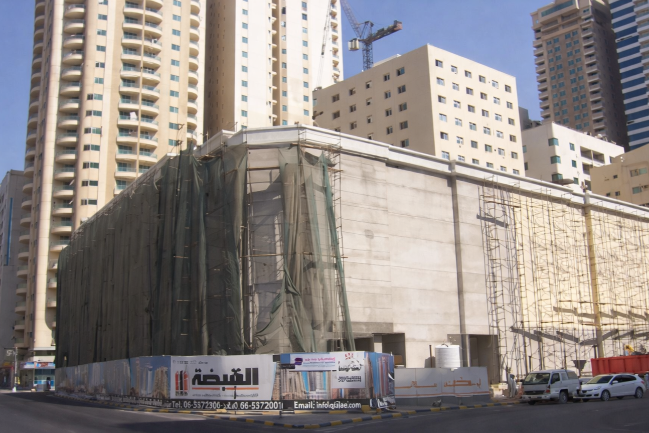 Construction site of a building in an urban area with high-rise buildings in the background, scaffolding, and a crane, with a fence displaying advertisements and contact information.