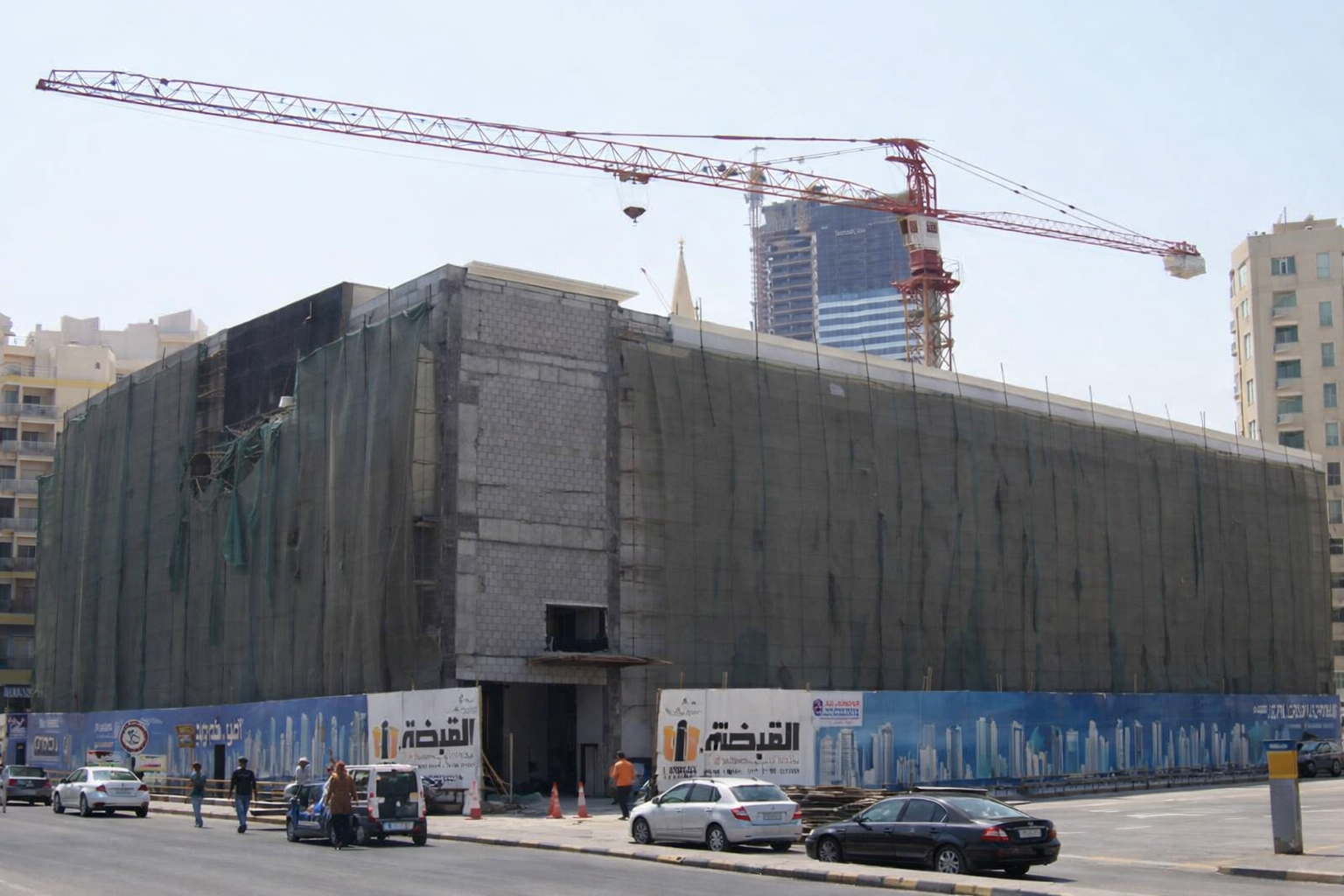 Construction site of a building with a large crane overhead, covered with green safety netting, in an urban area with high-rise buildings behind.