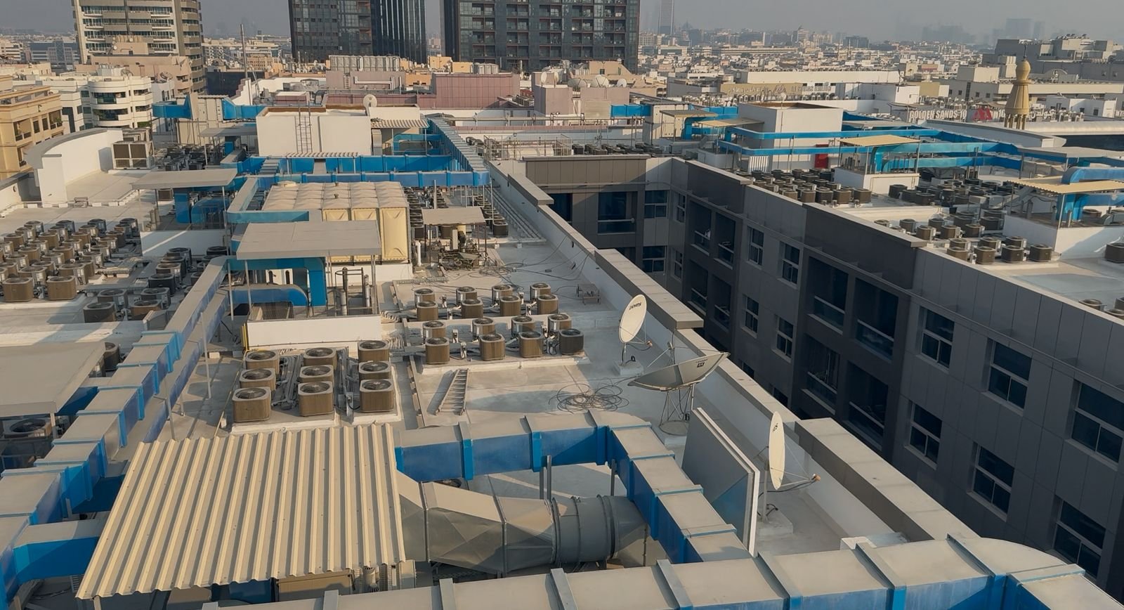 View of a building roof with HVAC units, satellite dishes, and blue ventilation ducts, in an urban cityscape with tall buildings in the background.