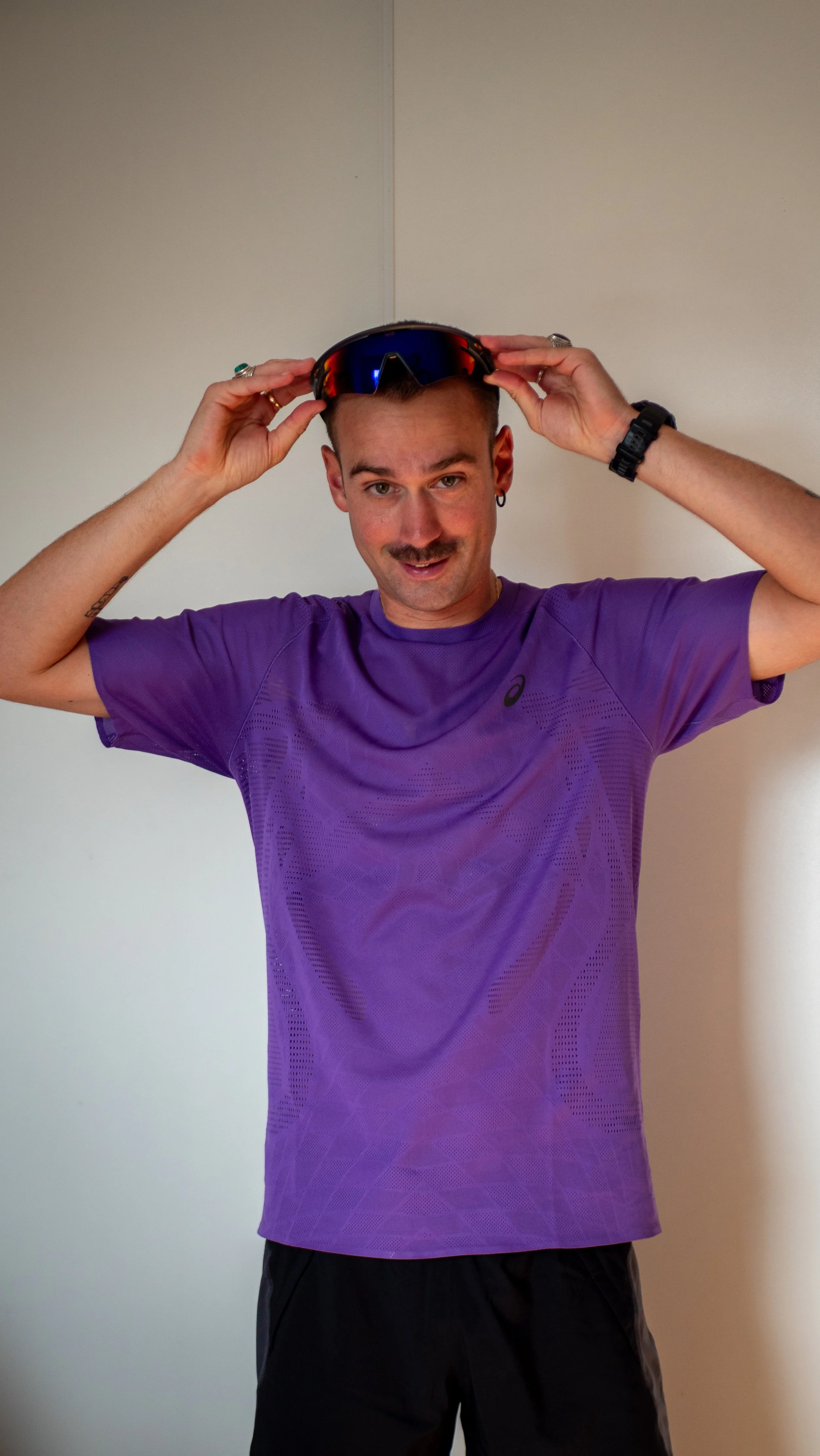 Un homme souriant portant un t-shirt violet, tenant des lunettes de soleil sur sa tête, avec un fond blanc.