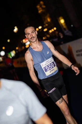 Un homme vêtu d'un maillot de course bleu et d'un court pantalon noir, portant un numéro de participant 559, participe à une course ou un marathon en soirée, avec des lumières de rue en arrière-plan.