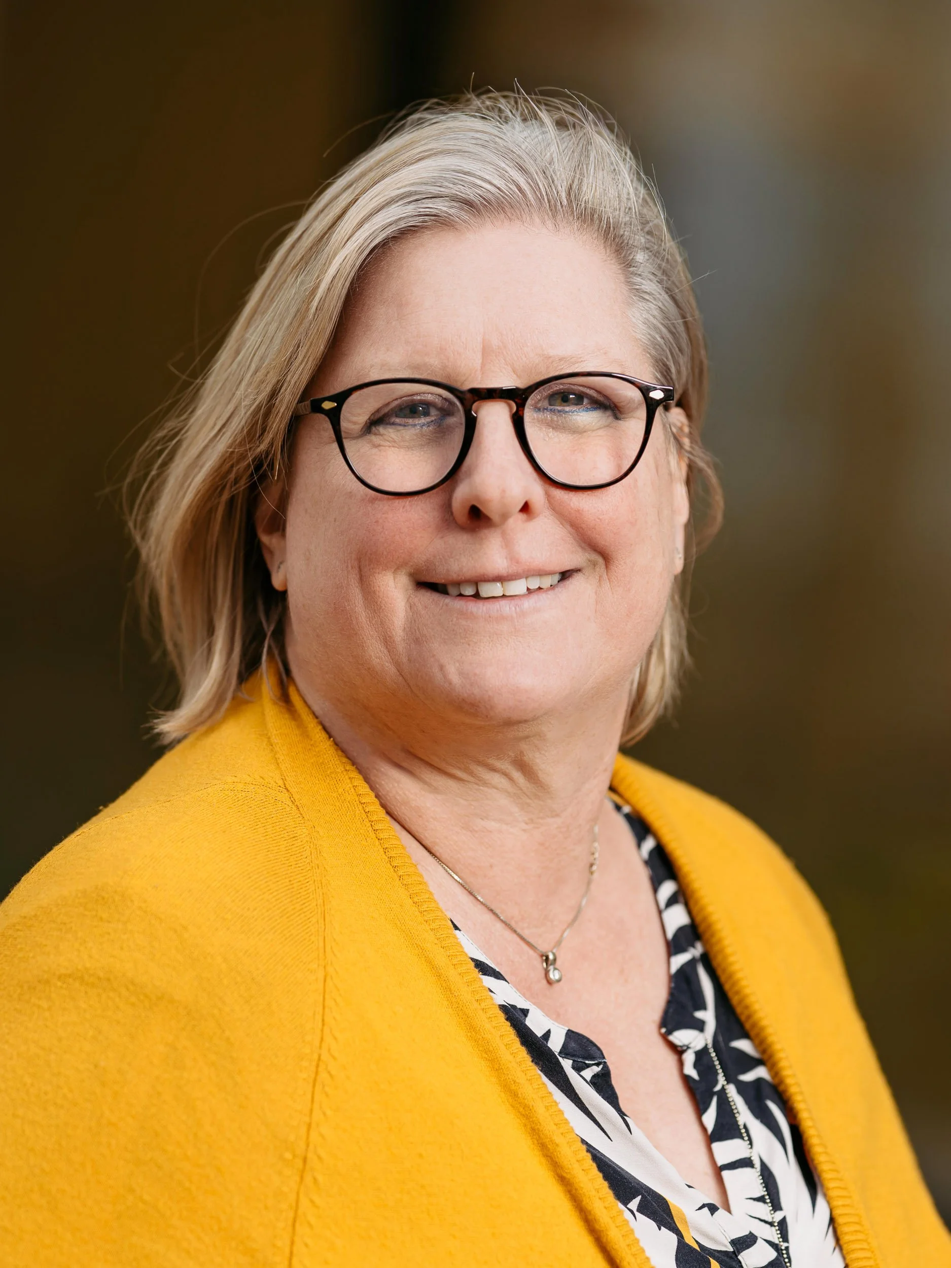 Porträt einer Frau mit kurzen, grauen Haaren, schwarzen Brillengläsern, gelber Jacke und schwarzen und weißen Mustermotiv. Sie lächelt und steht vor einem unscharfen, natürlichen Hintergrund.