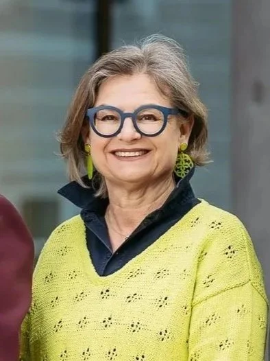 Eine ältere Frau mit kurzen grauen Haaren, blauer Brille, grünen Ohrringen und einem gelben Pullover, die lächelnd in die Kamera schaut.
