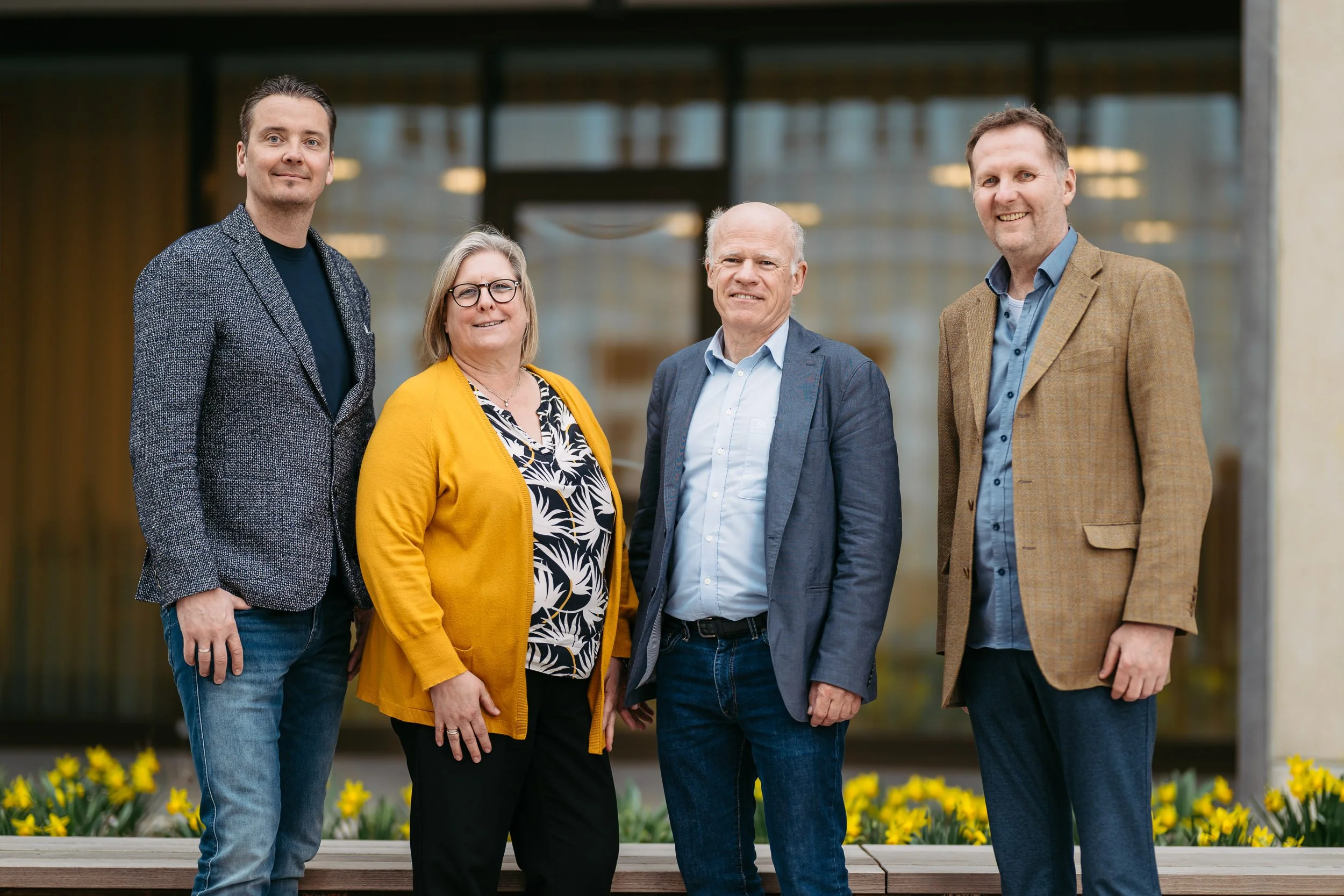 Fünf Personen stehen vor einem modernen Gebäude mit großen Fenstern, die im Hintergrund zu sehen sind, und einem Vorgarten mit gelben Blumen.