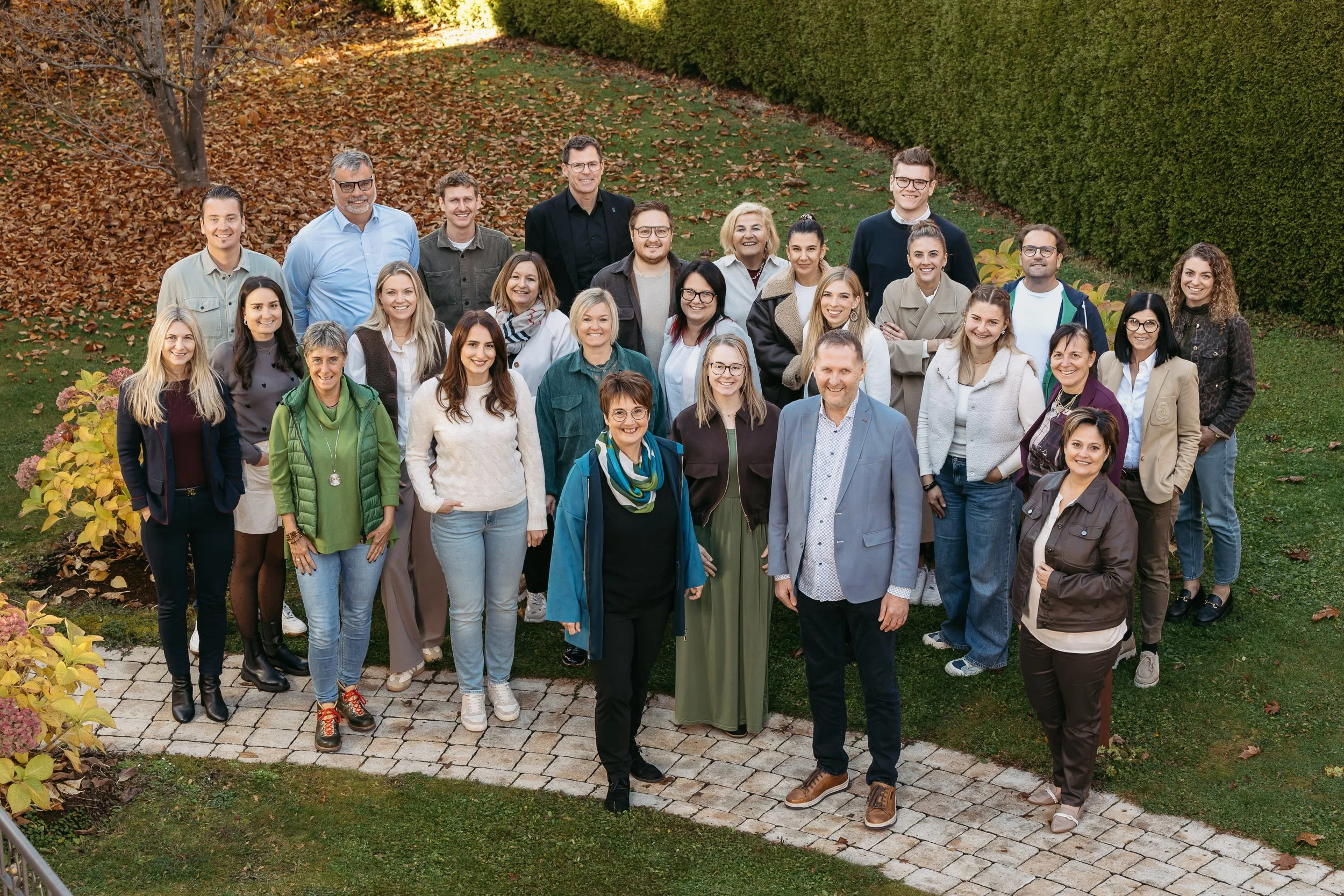 Gruppenfoto einer großen Gruppe von Menschen im Freien, im Herbst, auf einer gepflasterten Terrasse, mit Bäumen und einer Hecke im Hintergrund.