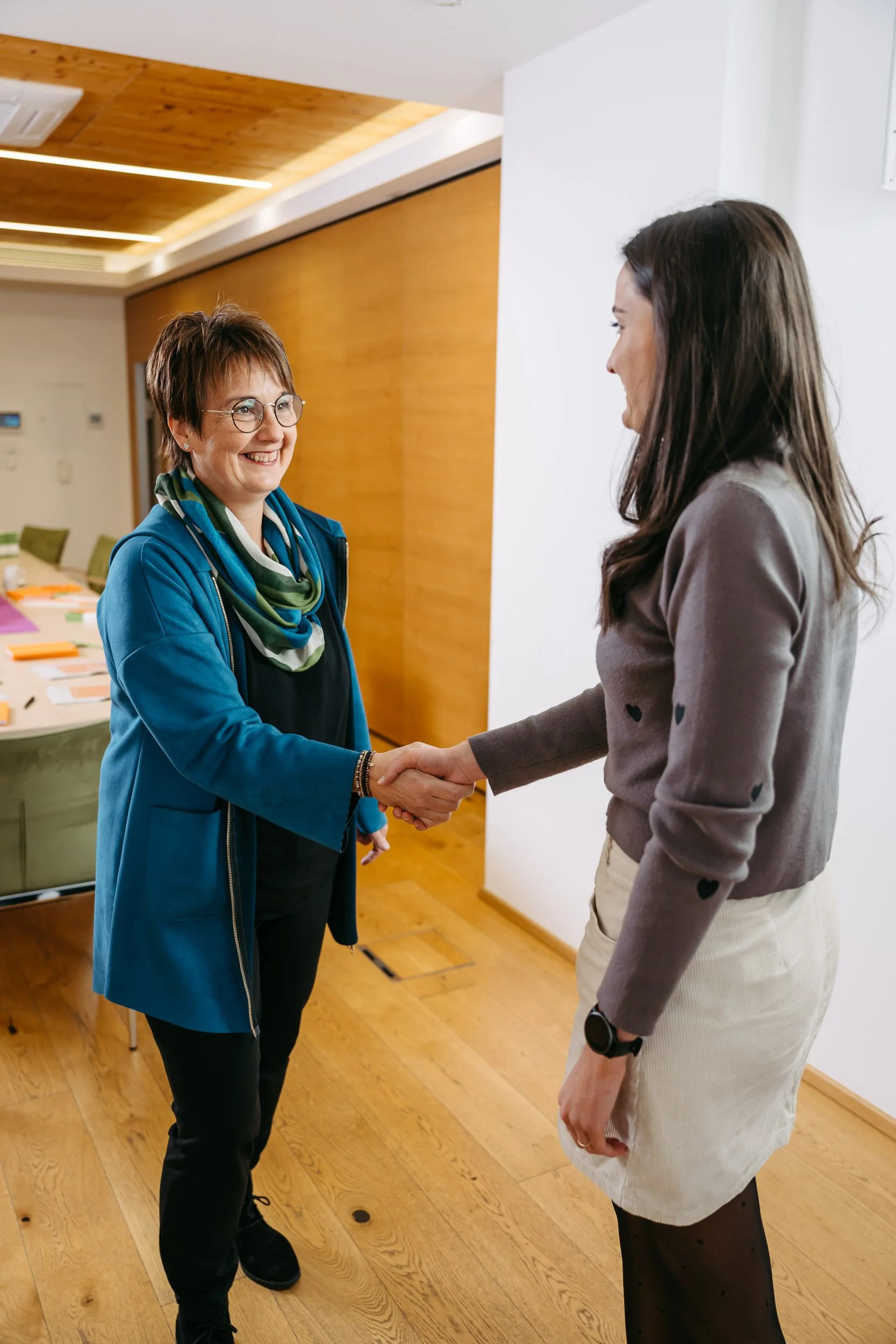 Zwei Frauen schütteln sich die Hand in einem modernen Raum mit Holzfußboden und hellem Holzdecke.