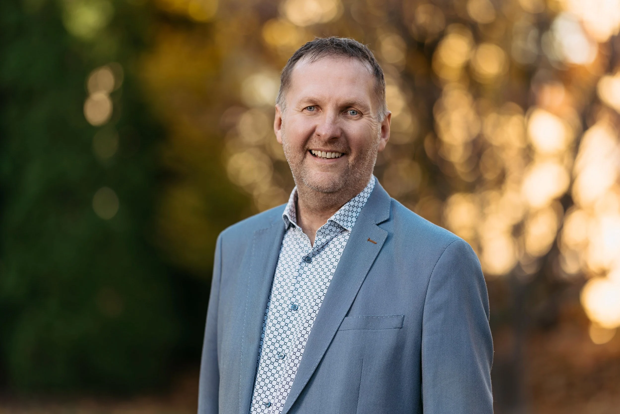 Ein lachender Mann in einem blauen Anzug vor einem herbstlichen Hintergrund mit warmem Licht.
