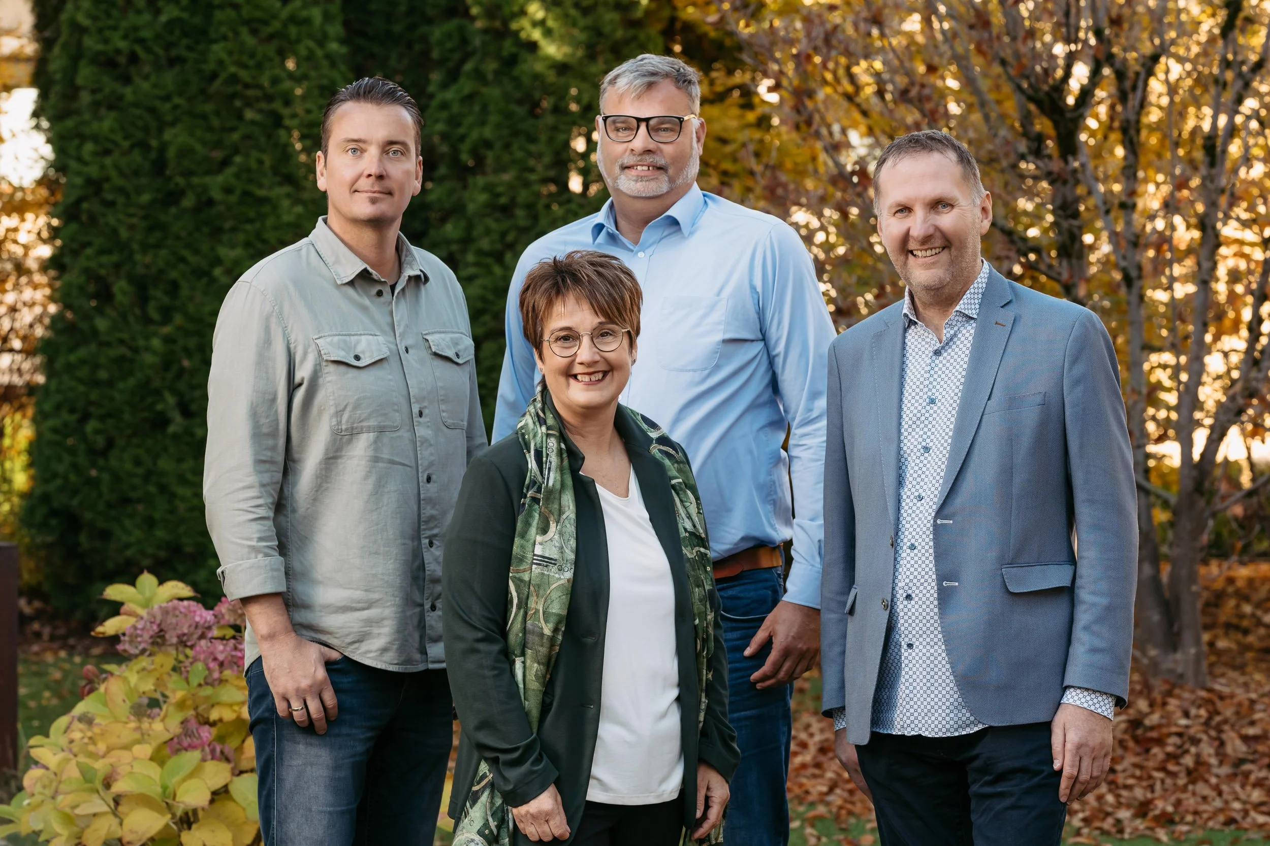 Fünf Menschen stehen im Garten mit Herbstlaub und Bäumen mit Gold- und Rottönen im Hintergrund, sie lächeln und posieren für ein Gruppenfoto.