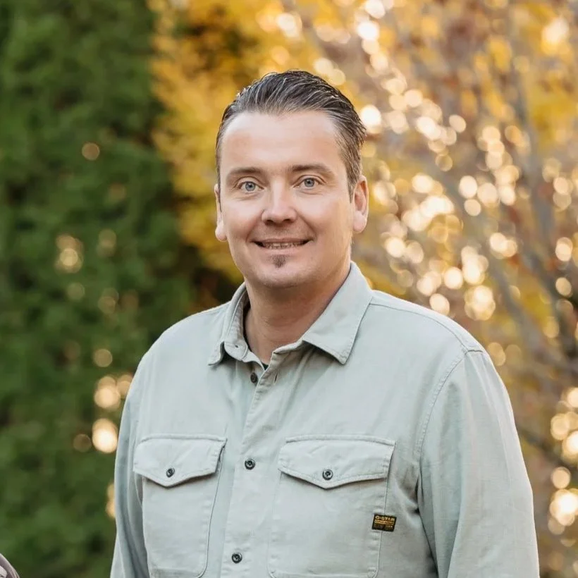 Ein Mann mit dunklen, nach hinten frisurierten Haaren, trägt ein hellgraues Hemd, steht im Freien bei Sonnenuntergang mit Baumlaub im Hintergrund.