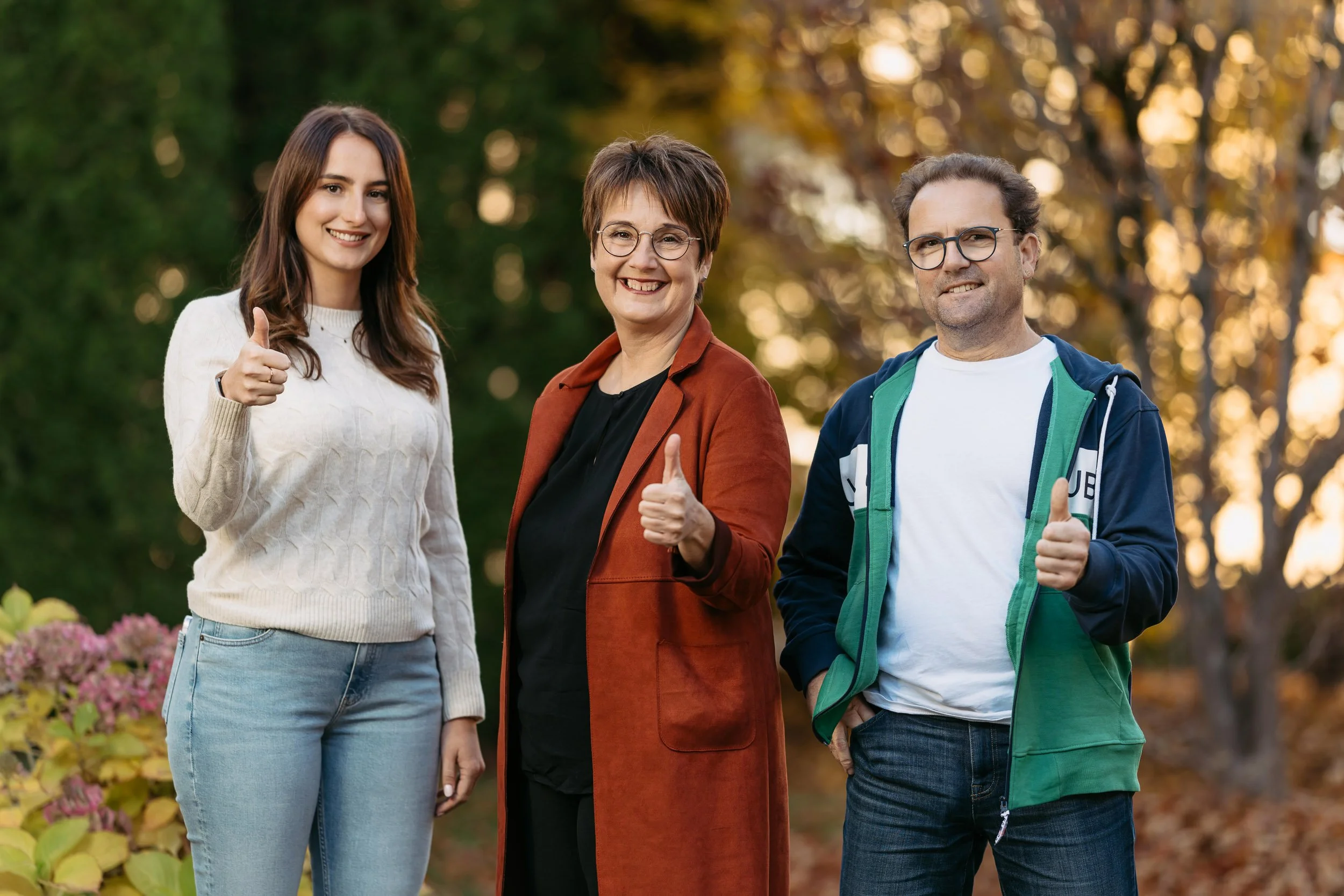 Drei Menschen stehen im Freien, lächelnd und geben Daumen hoch, bei herbstlichem Hintergrund mit Bäumen und Blumen.