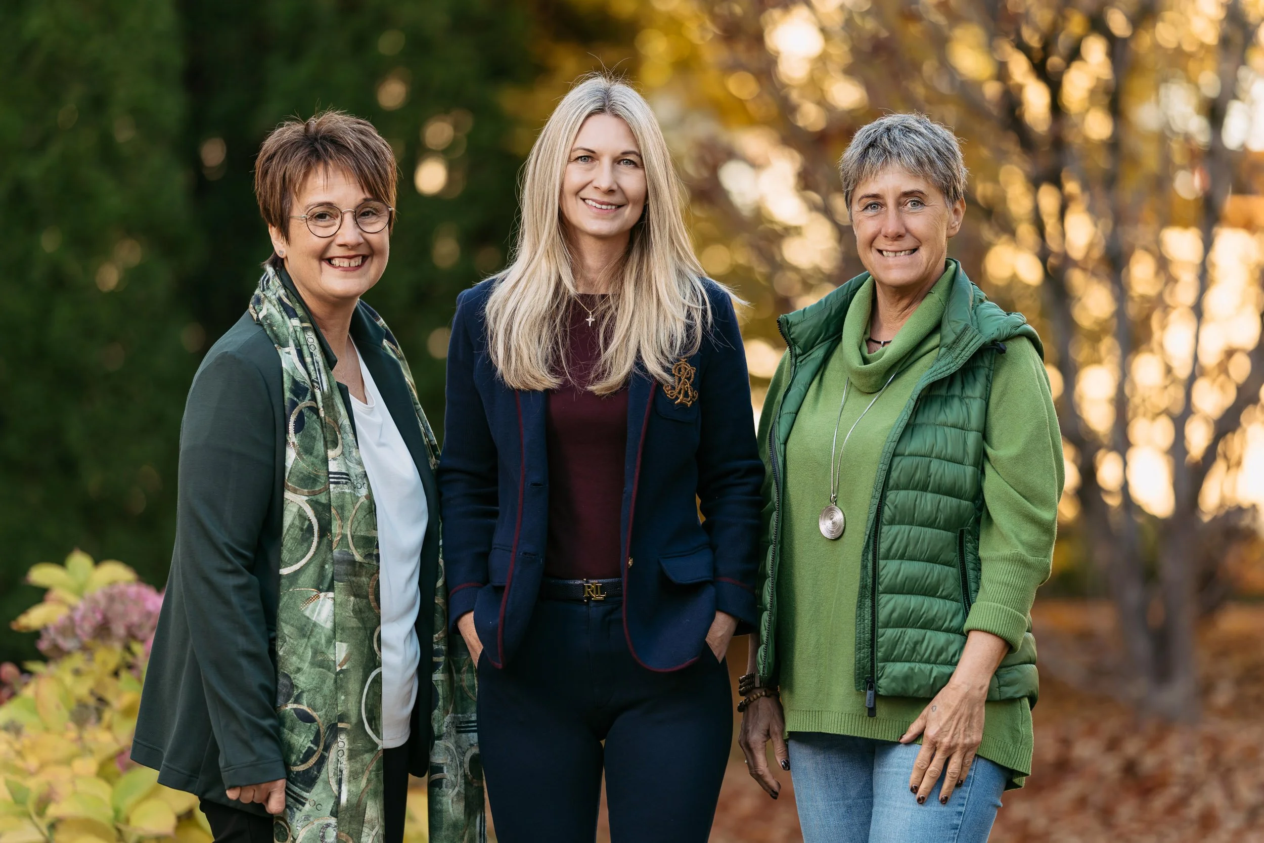 Drei Frauen stehen im Freien vor Baum mit herbstlichen Blättern, lächeln in die Kamera, tragen unterschiedliche Kleidung, eine trägt eine grüne Weste, eine trägt eine dunkle Jacke, die andere trägt eine Bluse und einen Schal.