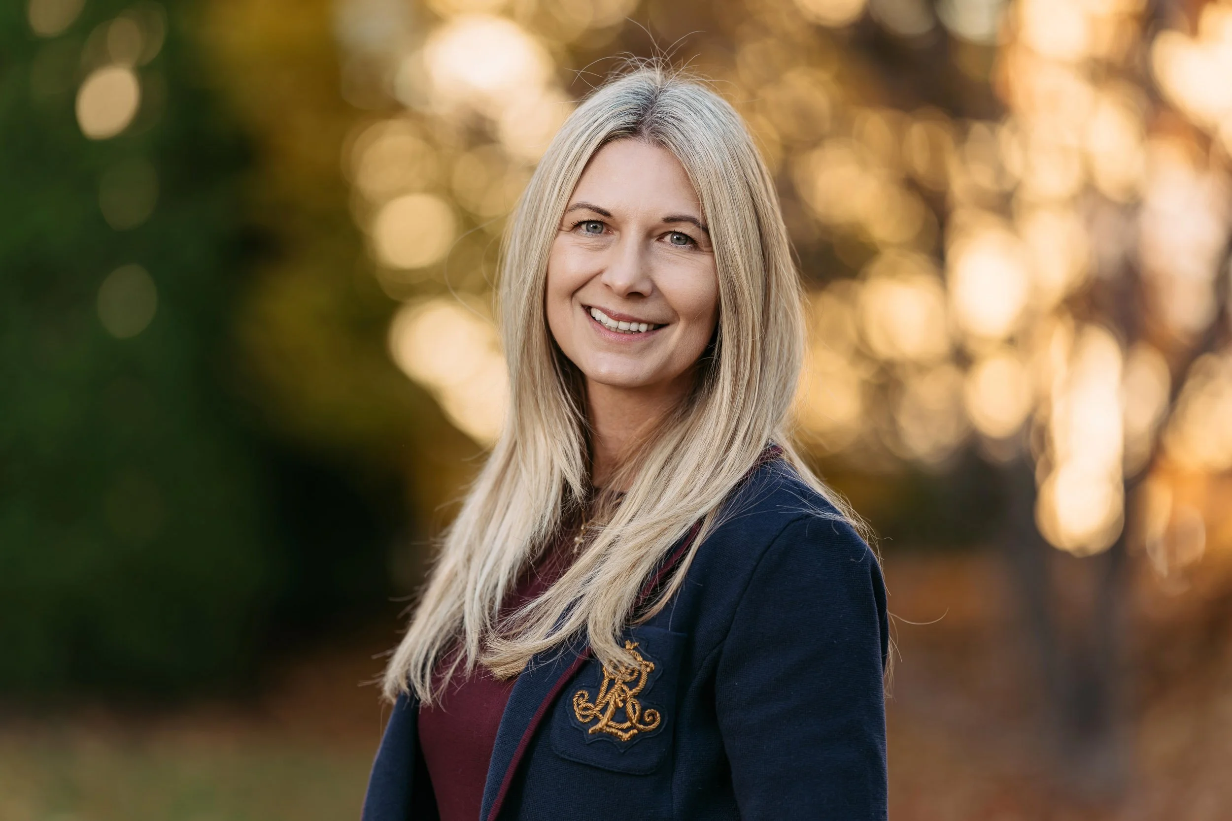 Eine lächelnde blonde Frau im Freien bei Sonnenuntergang, im Hintergrund bunte Herbstbäume.