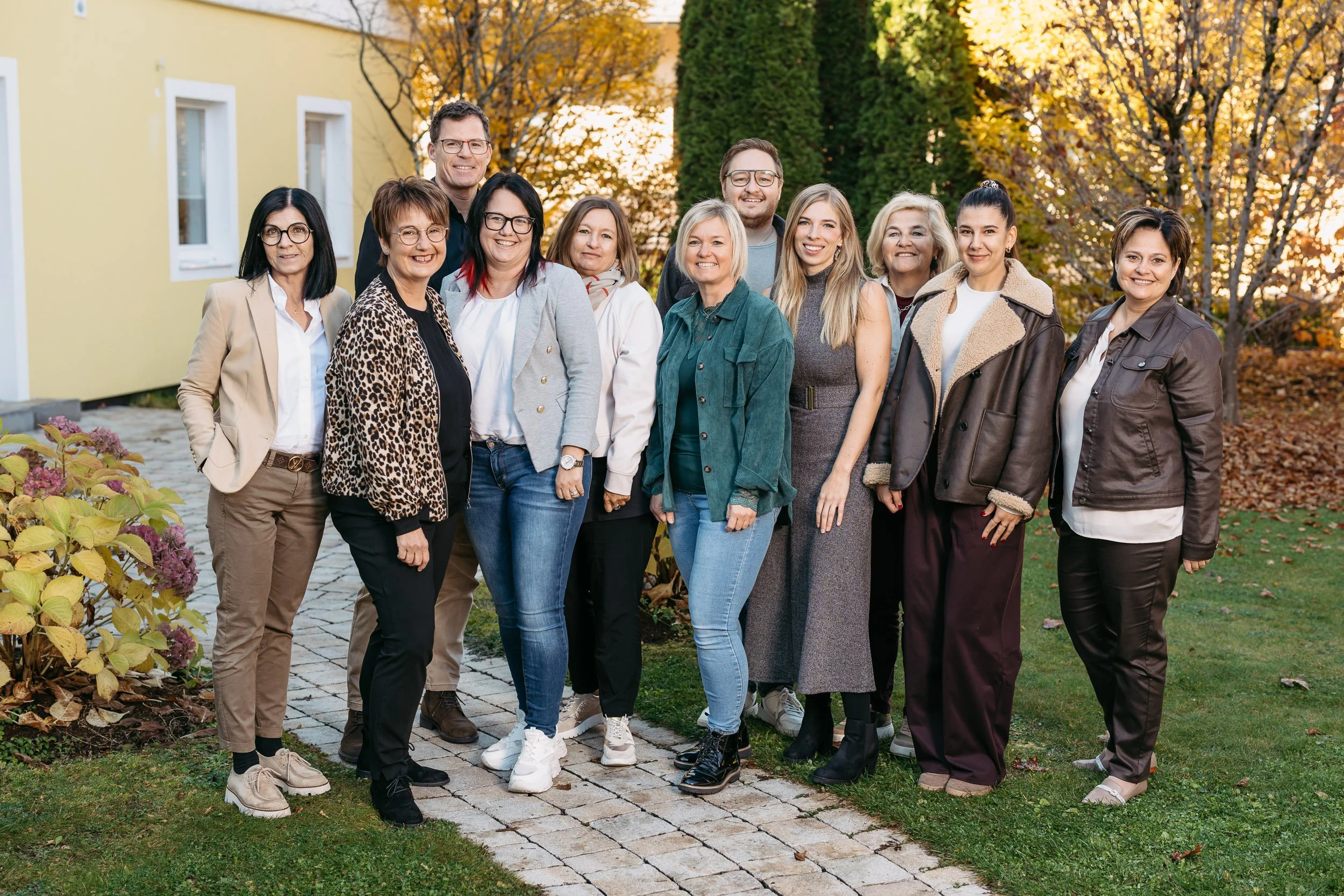 Gruppe von zwölf Menschen, vier Männer und acht Frauen, stehen draußen im Herbst vor einem gelben Haus und bunten Bäumen, lächelnd und in lockerer Kleidung.